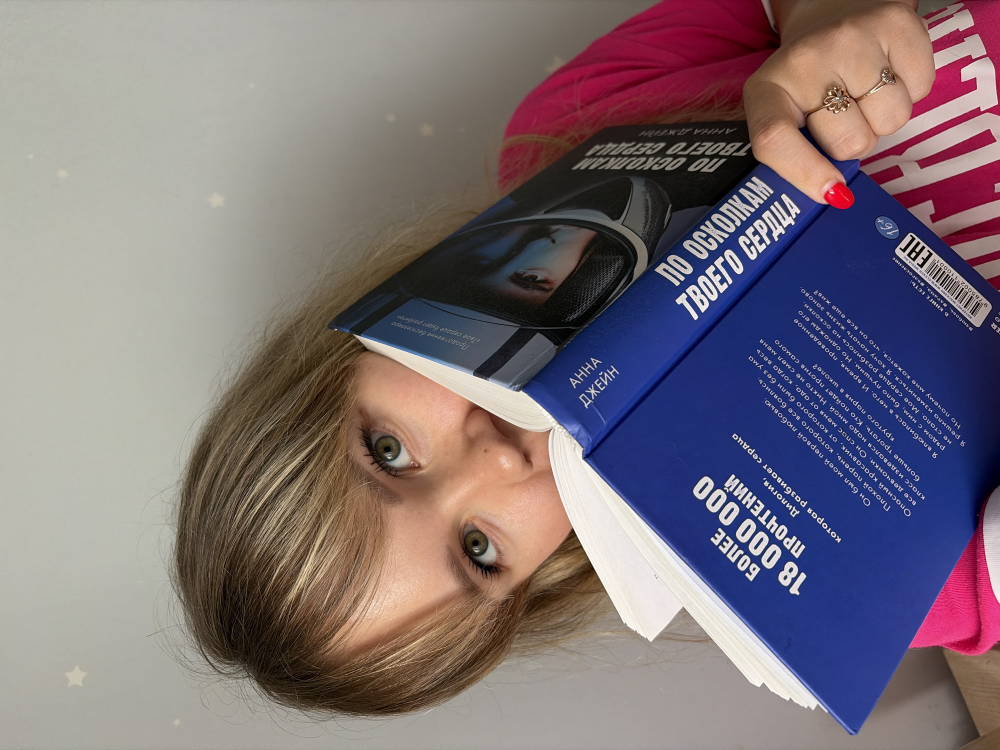 Here is a forensic description of the image:
**People:** A Caucasian woman with blonde hair and light green eyes is visible. She is wearing a pink and white striped shirt. Her left hand is visible, adorned with two gold rings and a red nail polish on the index finger.
**Subjects:** The main subject is a book. The title is "ПО ОСКОЛКАМ ТВОЕГО СЕРДЦА" (translated: "By the Shards of Your Heart"). The author is listed as "АННА ДЖЕЙН" (Anna Jane). The book has a blue cover with white text and another cover featuring a person in a spacesuit.
**Objects:** Besides the book, a wall with small star decals is visible in the background.
**Actions:** The woman is holding the book partly over her face, in a way that covers her forehead.
**Interactions:** The woman is interacting with the book by holding it.
**Environment:** The background is a room with a light-colored wall and star decorations. Based on the Cyrillic text on the book and the information provided, the image is likely taken in Russia, though the city of Chaikovsky cannot be identified from the details available in the image.