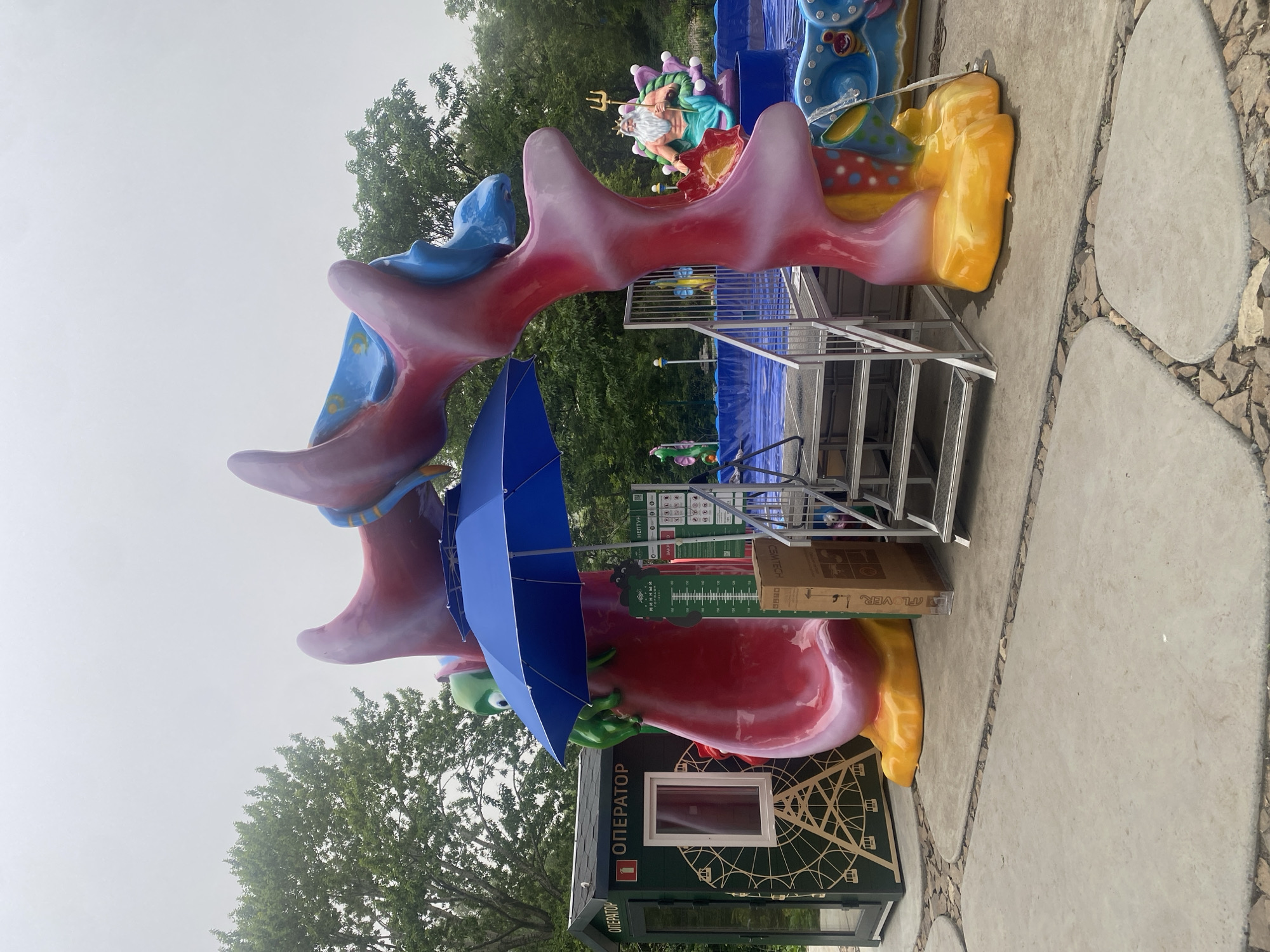 The image depicts a colorful amusement park ride with an underwater theme, possibly a water play area or a themed slide, set against a backdrop of trees and an overcast sky. The ride features vibrant, sculptural elements resembling coral and sea creatures. In the foreground, a large, red and pink coral-like structure forms an archway. A green cartoonish sea creature is visible on the left side of the arch, near a blue umbrella. Metal stairs lead up to the ride's platform. To the left of the ride is a small booth labeled "ОПЕРАТОР" (Operator) with a graphic of a Ferris wheel on its side. In the background, a pool area with blue lining is visible, and a statue of a bearded man with a trident, resembling Neptune, is perched on a purple shell. The ground is paved with concrete slabs and small stones.