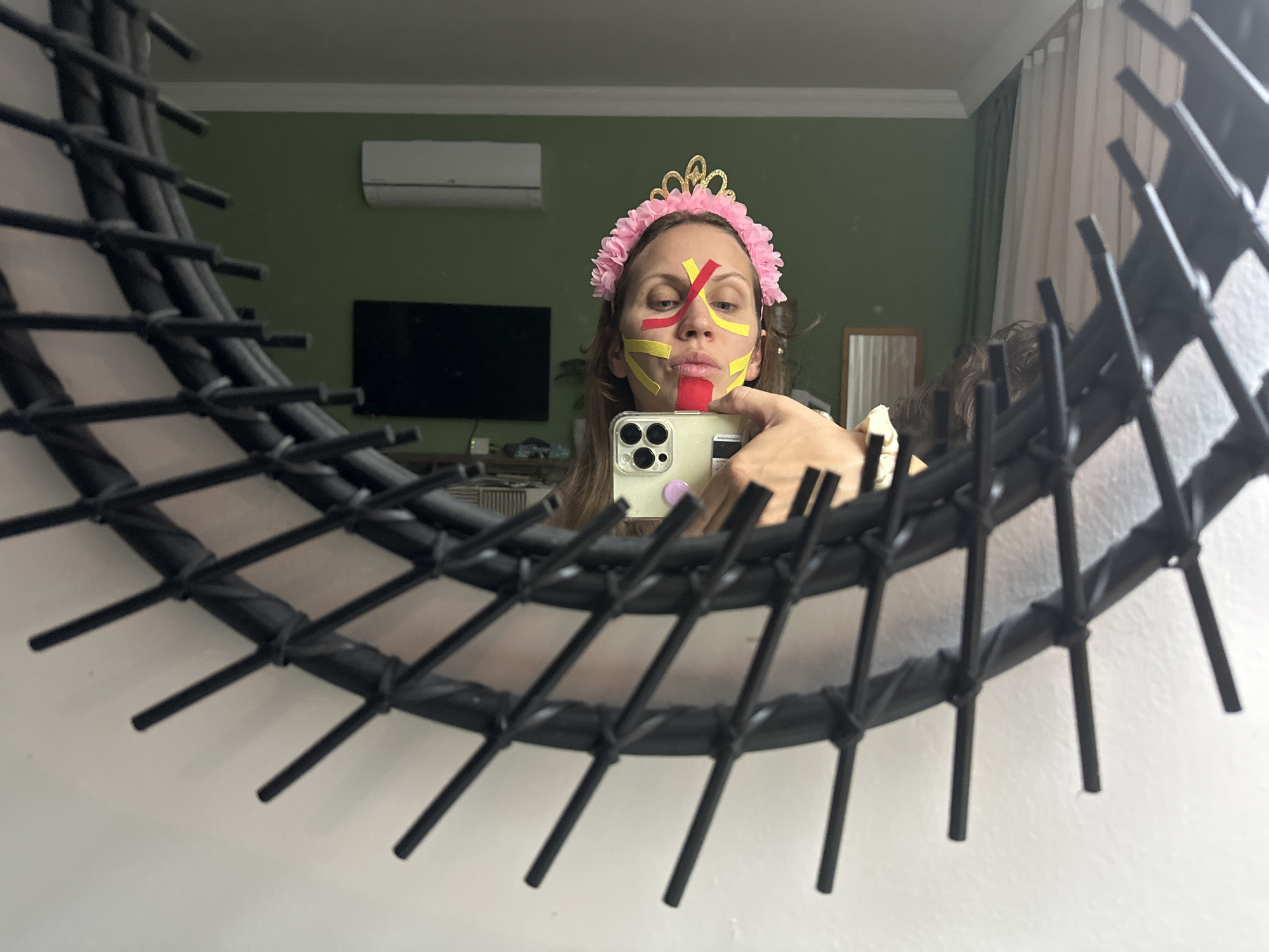 A woman is captured in a mirror selfie, her reflection showing her face adorned with red and yellow adhesive tape, forming a distinctive, flag-like pattern across her nose, cheeks, and upper lip. She wears a pink floral headband with a golden tiara. Her brown hair frames her face as she holds an iPhone, visible in the lower part of the frame, to take the picture.
The mirror itself has a prominent, intricate black woven or lattice-like frame, which partially obscures the edges of the reflection, particularly on the left and bottom right. The background, reflected in the mirror, reveals an indoor setting, likely in Fethiye, Turkey. A green wall with white crown molding is visible, along with a white air conditioning unit mounted high on the wall. Below it, a black television screen is partially seen. To the right, white curtains and a glimpse of another smaller mirror contribute to the room's decor. The woman's expression is neutral, suggesting focus on taking the selfie while showcasing her festive face paint and headwear. The lighting indicates an indoor scene, with no specific time of day discernible. No explicit text is visible in the image.