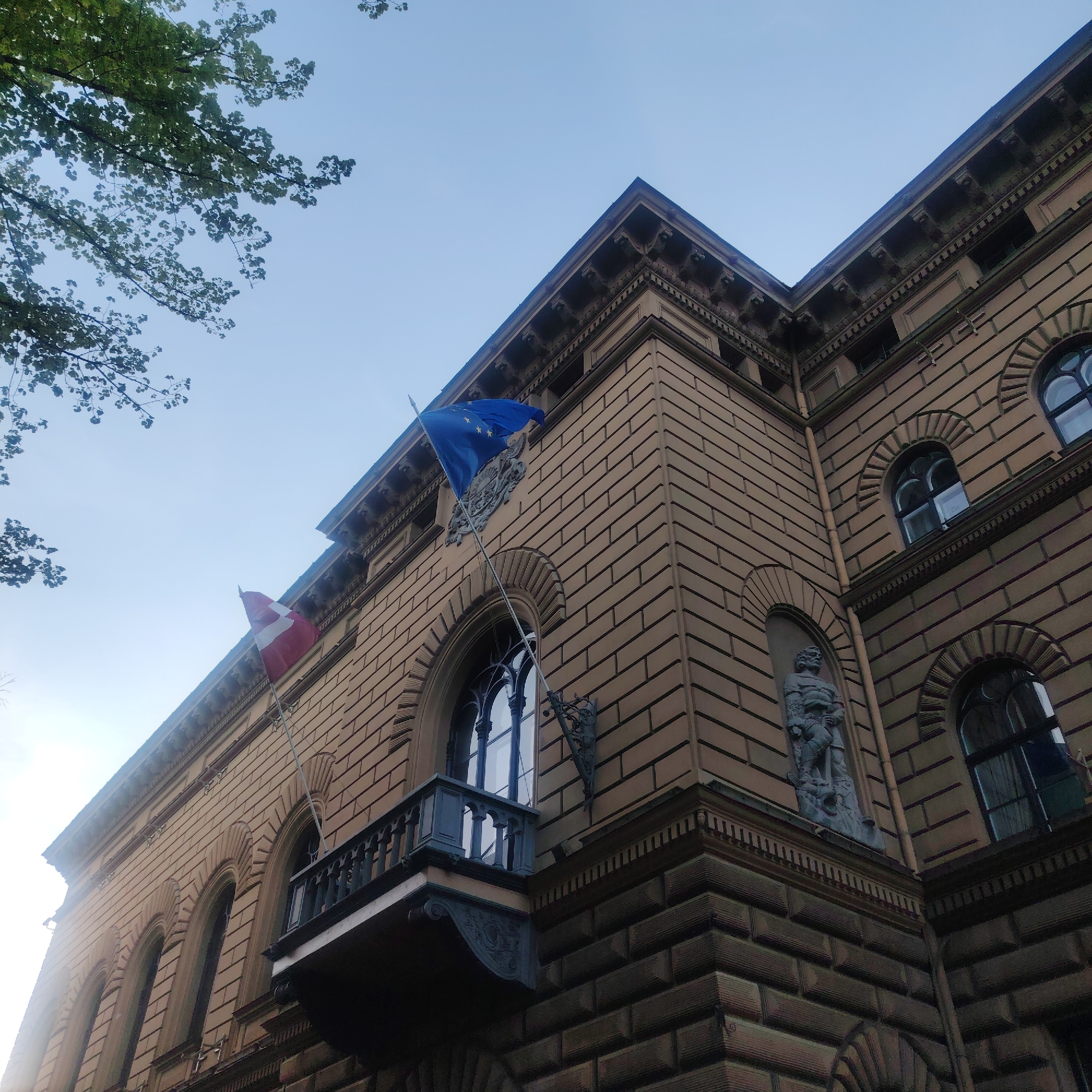 The image shows a low-angle view of a building in Riga, Latvia. The building has a beige facade with a textured block pattern. Arched windows with dark frames are visible along the facade. A small balcony with dark railings projects from one section of the building. Two flags are flying from the building; one appears to be the flag of Latvia, and the other is the flag of the European Union. There is a statue of a person in armor located above one of the windows. Parts of a tree are visible in the upper left corner of the frame, and the sky is blue.