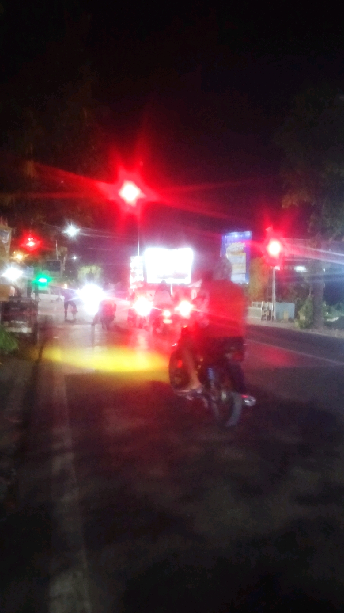 The image depicts a nighttime scene on a street in Bojonegoro, Indonesia. Several motorcycles are visible, with one in the foreground partially obscuring the view of other vehicles and pedestrians. Red traffic lights are illuminated, casting a strong red glow on the surroundings. The street appears wet, possibly due to rain, reflecting the lights from traffic signals and vehicle headlights. The environment suggests a typical urban setting with buildings and trees lining the road, though details are blurred due to the low light conditions and motion. The overall impression is one of a busy intersection at night, with the red traffic lights creating a dramatic and somewhat abstract atmosphere.