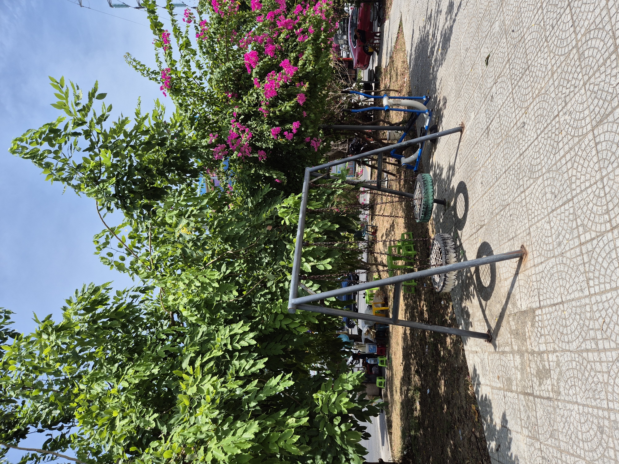 A children's swing set with two tire seats is positioned on a tiled walkway under the shade of large trees. One tree has dense green foliage, while another displays a vibrant display of pink flowers. In the background, a red car is parked near a sidewalk. The scene appears to be an outdoor public space, possibly a park or playground, located in the city of Đà Nẵng, Vietnam, given the context provided.