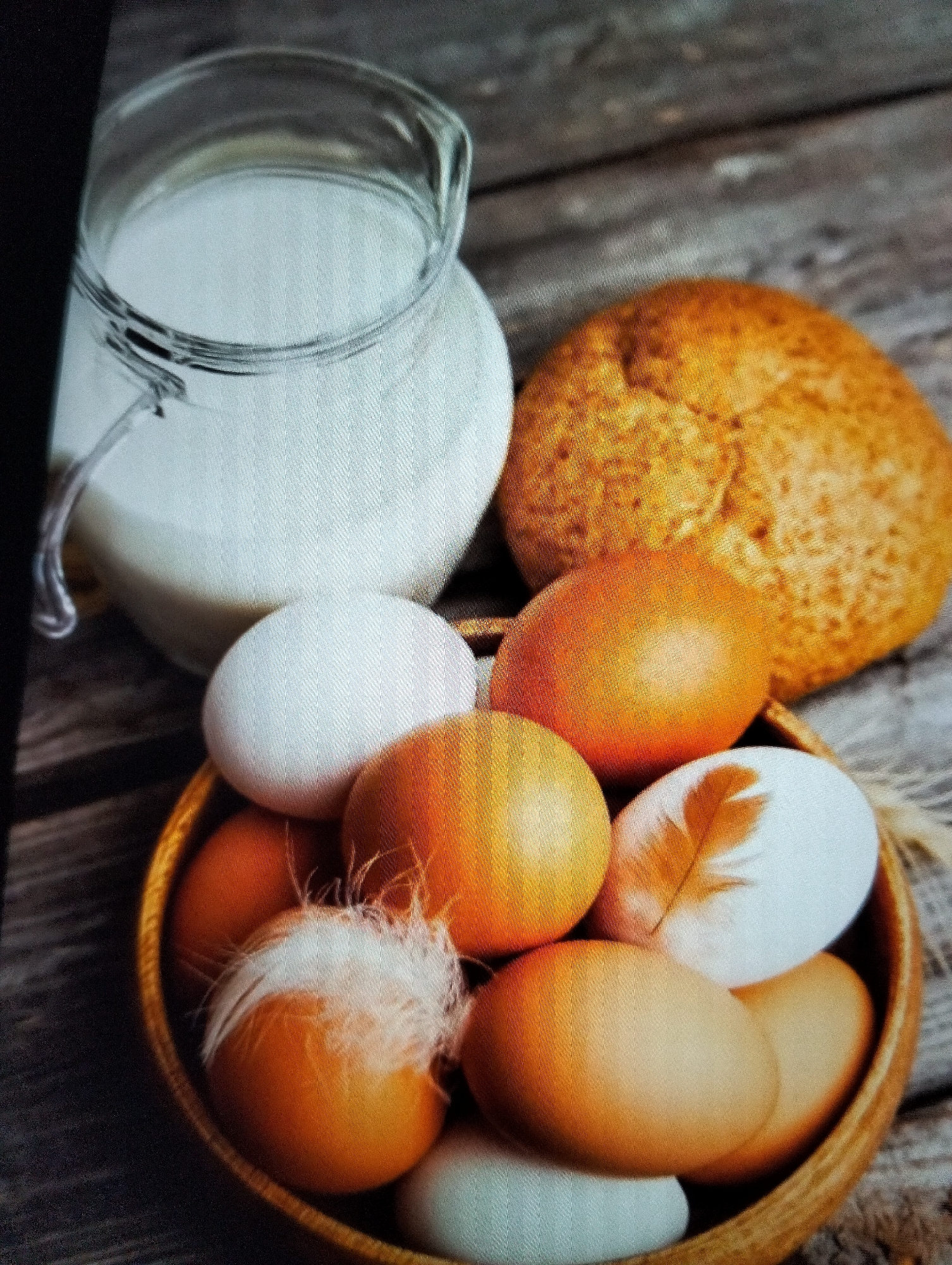 An overhead shot shows a glass pitcher filled with milk, a golden-brown bread roll, and a wooden bowl containing several eggs, some with feathers. The scene is set on a rustic wooden surface. The location is not specified within the image or the provided context.