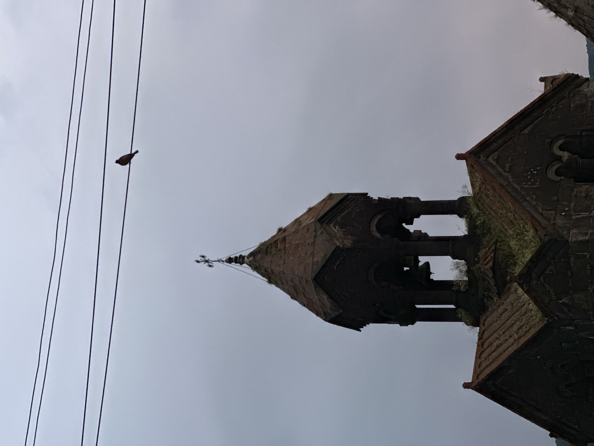 In Haghpat, Armenia, a small bird is perched on a horizontal wire. Below the bird and wire, the conical, stone roof of an ancient church structure is visible, topped with a cross. The sky is overcast and grey.