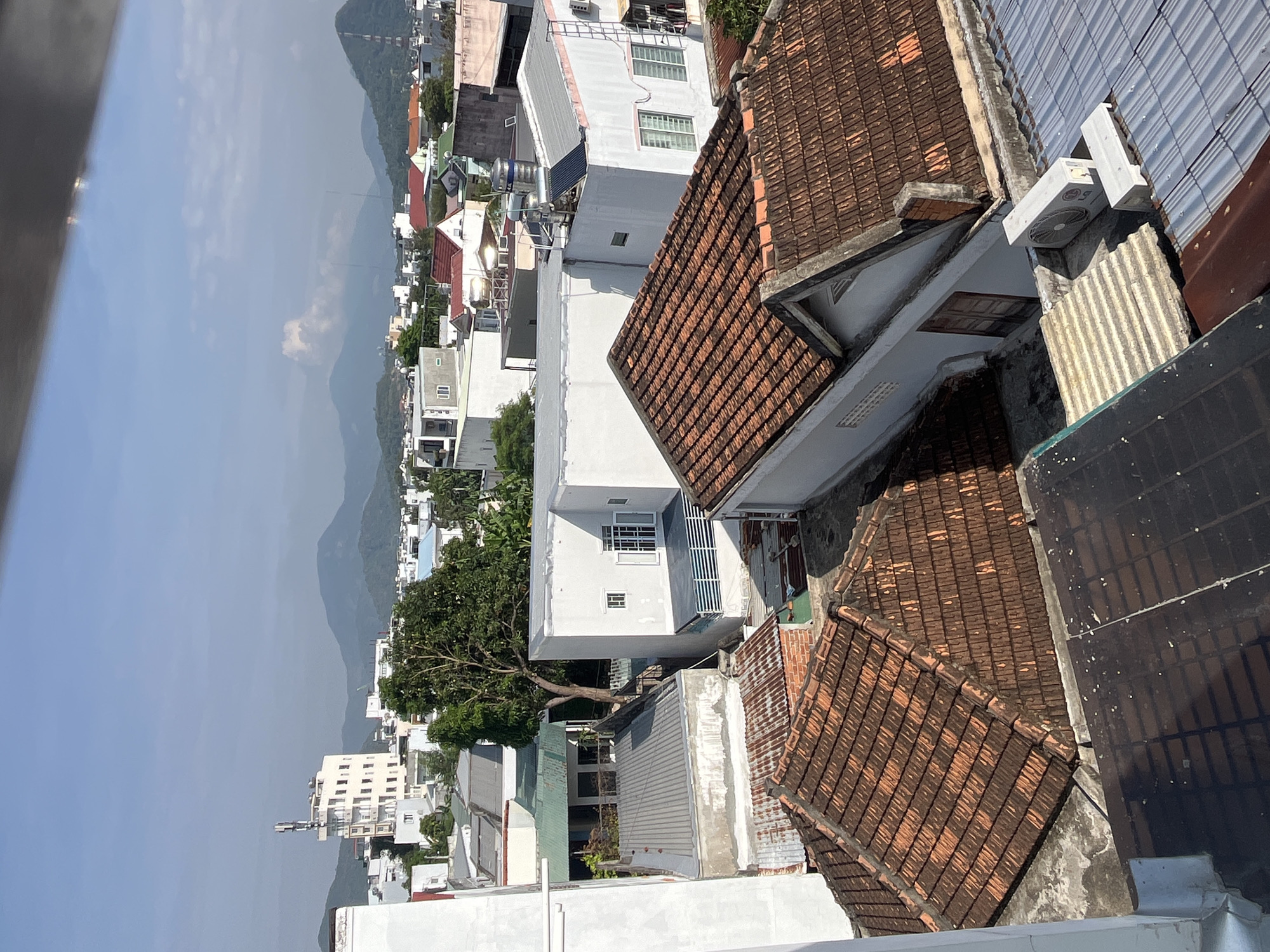 This image depicts a high-angle view of a residential area in Bac Nha Trang Ward, Vietnam, showing a mix of buildings with red tile roofs and white facades. In the background are distant mountains obscured by a slightly hazy sky. The foreground consists of rooftops and building structures with various materials, including corrugated metal and painted concrete. No people or animals are visible. The composition emphasizes the density of the urban landscape set against the backdrop of the natural environment.