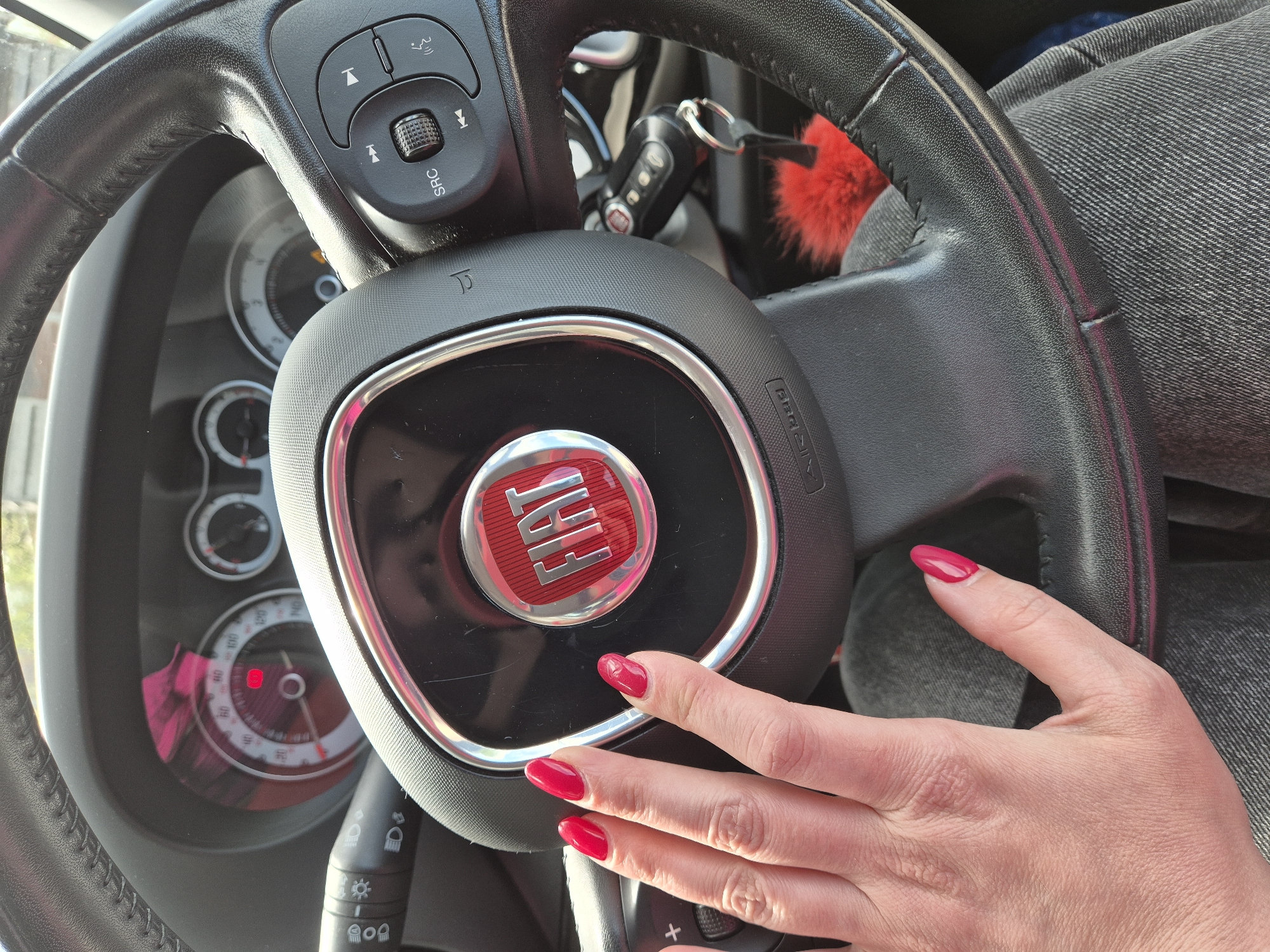 The image depicts the interior of a Fiat car. A person's hand with red nail polish rests on the steering wheel, near the Fiat logo. The steering wheel features buttons for audio and phone control. Car keys with a red fluffy keychain are visible on the steering column. The dashboard and speedometer are partially visible behind the steering wheel. The person is wearing grey jeans. The location is not discernable from the image, therefore it is not possible to determine if it is located in West Suffolk, United Kingdom.
