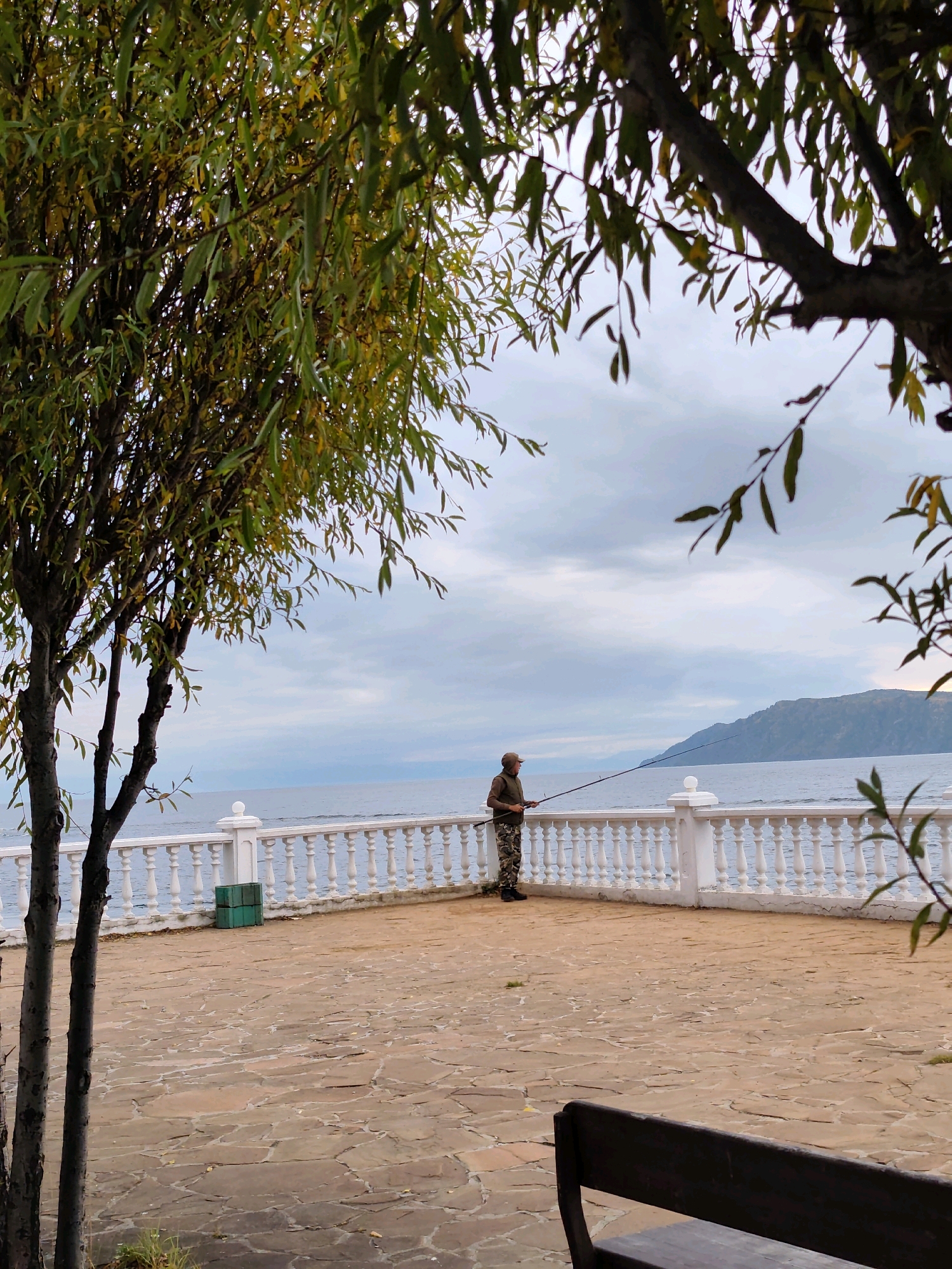 A person stands on a paved promenade next to a white balustrade, holding a fishing rod. The person is wearing camouflage pants and a dark jacket and cap. The promenade overlooks a body of water, possibly Lake Baikal, with a forested hillside in the distance. The sky is overcast. Trees with green and yellowing leaves frame the scene from the left and right. The promenade is paved with irregular stone tiles. In the background, to the left of the fishing person, are two green crates. A dark wooden bench is visible in the foreground, partially obscuring the view of the promenade. This scene is likely located in Listvyanka, Russia, a village on the shores of Lake Baikal.