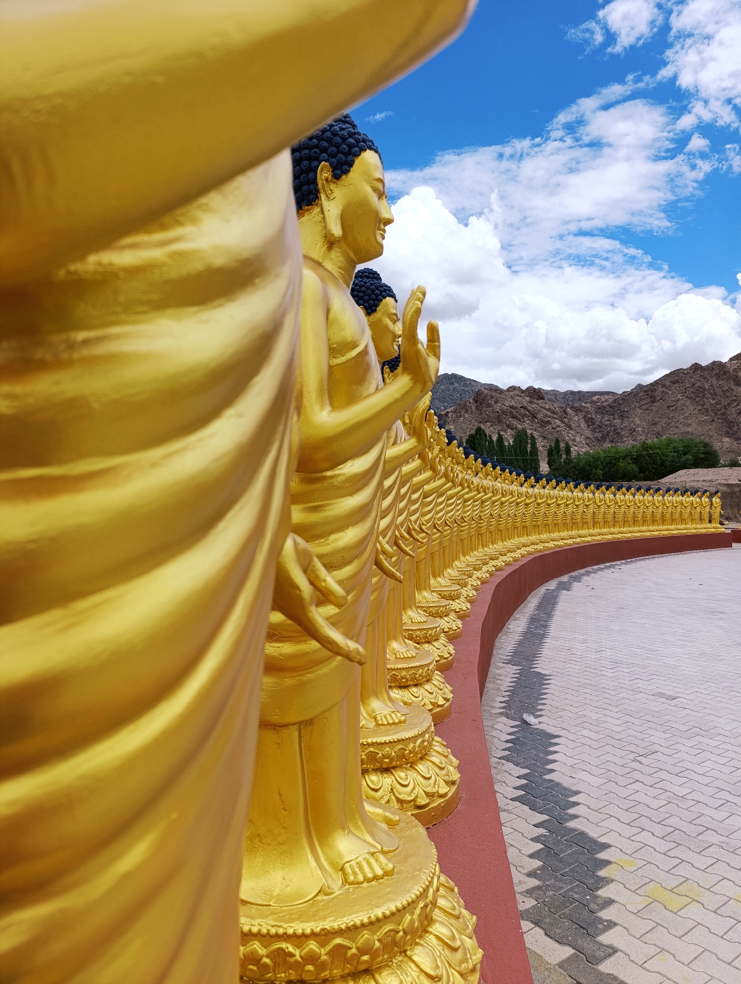 In Leh, India, a long, curving line of golden Buddha statues stands facing right, receding into the distance. The statues are arranged in a semi-circle with their backs to the viewer, each with a serene expression, dark blue hair, and draped robes. The foreground is dominated by the tightly cropped, golden form of a single Buddha statue, its draped robes rendered in striking detail and texture. The ground is paved with light-colored bricks arranged in a pattern, with a red border separating it from the statues. In the background, lush green trees line the base of rugged, brown mountains under a bright blue sky dotted with fluffy white clouds.