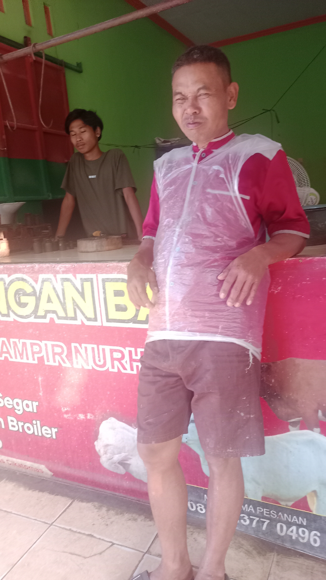 The image shows two men in front of a food stall in Karyamandala, Indonesia. The man in the foreground is an adult male with short hair, wearing a red short-sleeved shirt and brown shorts. He has a clear plastic apron over his shirt and is standing with his hands on his hips, facing the camera. The man in the background is younger, with dark, short hair, wearing a dark green t-shirt. He is standing behind a counter, with his forearms on the counter. The stall has a red and white sign with yellow lettering, which appears to be a menu or advertisement. The background is a green wall and a red and green structure, possibly a roof or awning. The floor is tiled.