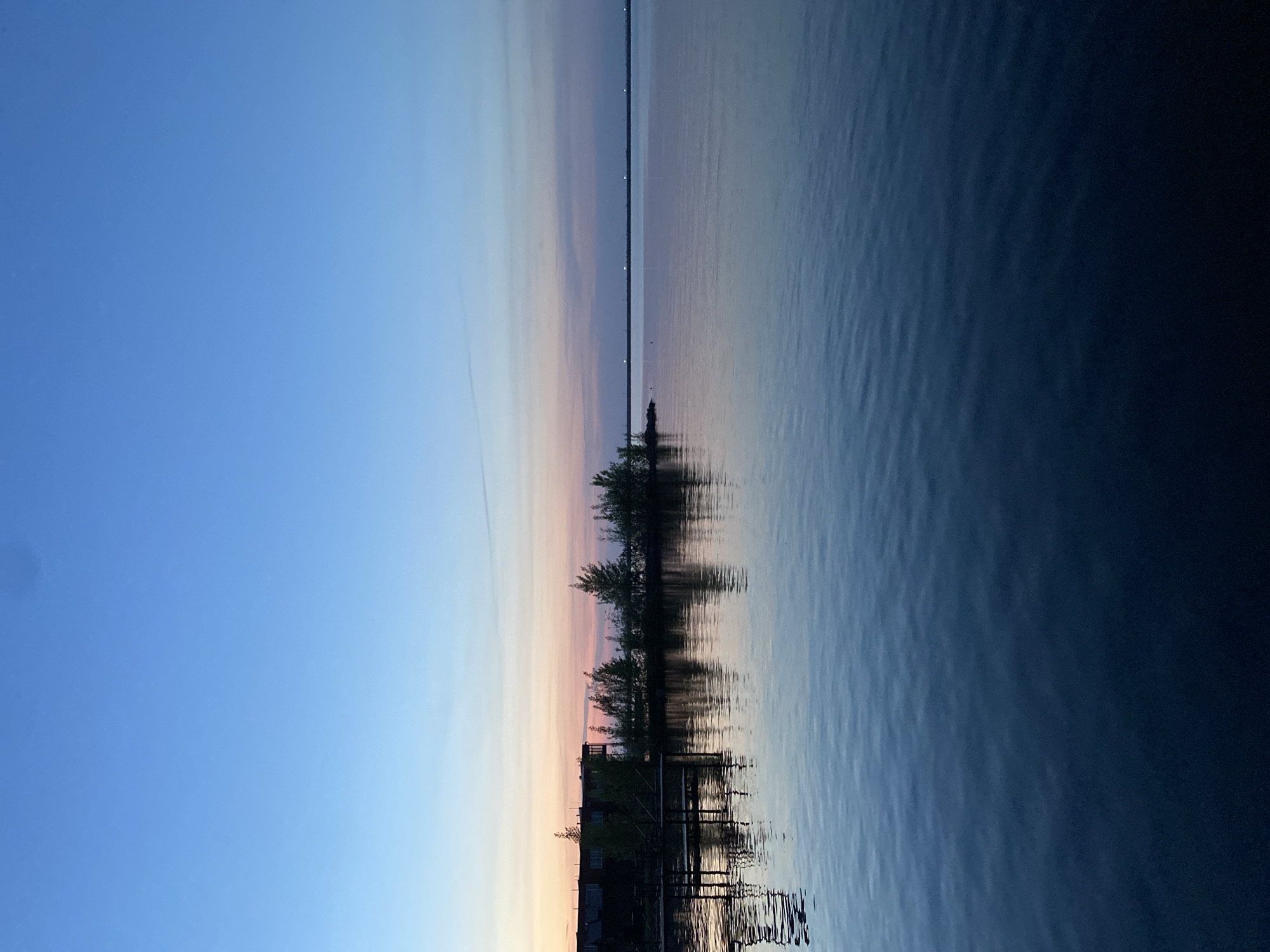 The image depicts a landscape at sunset or twilight in Chaikovsky, Russia. The sky transitions from a light blue on the left to hues of orange, pink, and purple towards the center horizon. A body of water occupies the right portion of the image with a dark blue tone. A treeline sits on the shoreline, reflecting in the water. Also visible are buildings and a structure that resembles a dock. A long strip of land or a structure runs horizontally across the middle ground of the image. The water surface has slight ripples, indicating calm conditions. The lighting suggests that the photo was taken during either dusk or dawn.