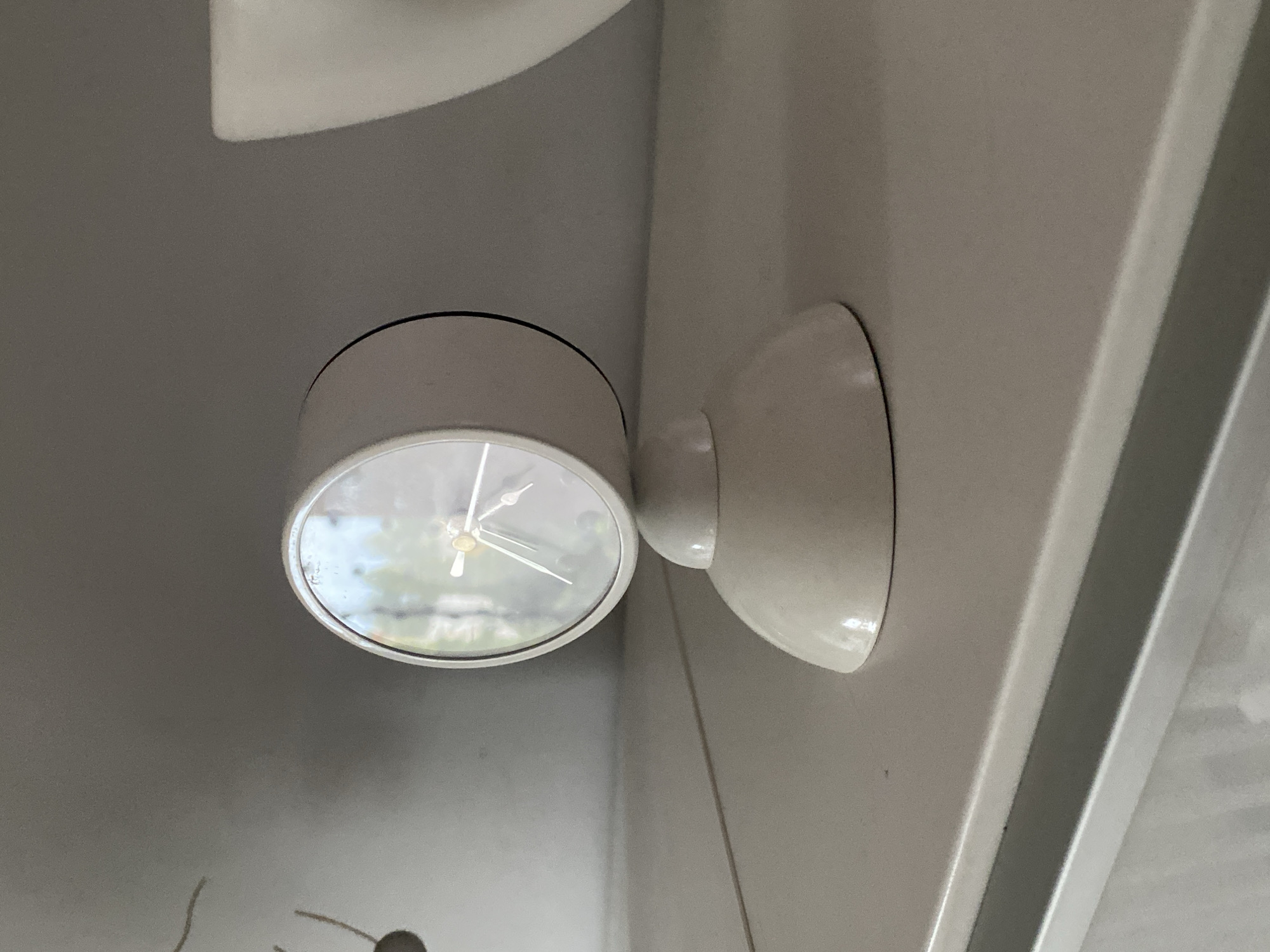 The image shows a close-up, eye-level shot of a white, retro-style alarm clock. The clock has a rounded base and a circular face with silver hands and Roman numerals. The face is reflective, showing a blurred reflection of trees and a building. The clock is positioned on a light-colored, flat surface, possibly a shelf or table. To the right of the clock, a portion of a white, curved object, possibly a vase or bowl, is visible. The background is a neutral, gray wall. The scene appears to be indoors, in a well-lit environment. The image does not contain any people or indicate a specific location within the city of Wrocław, Poland.