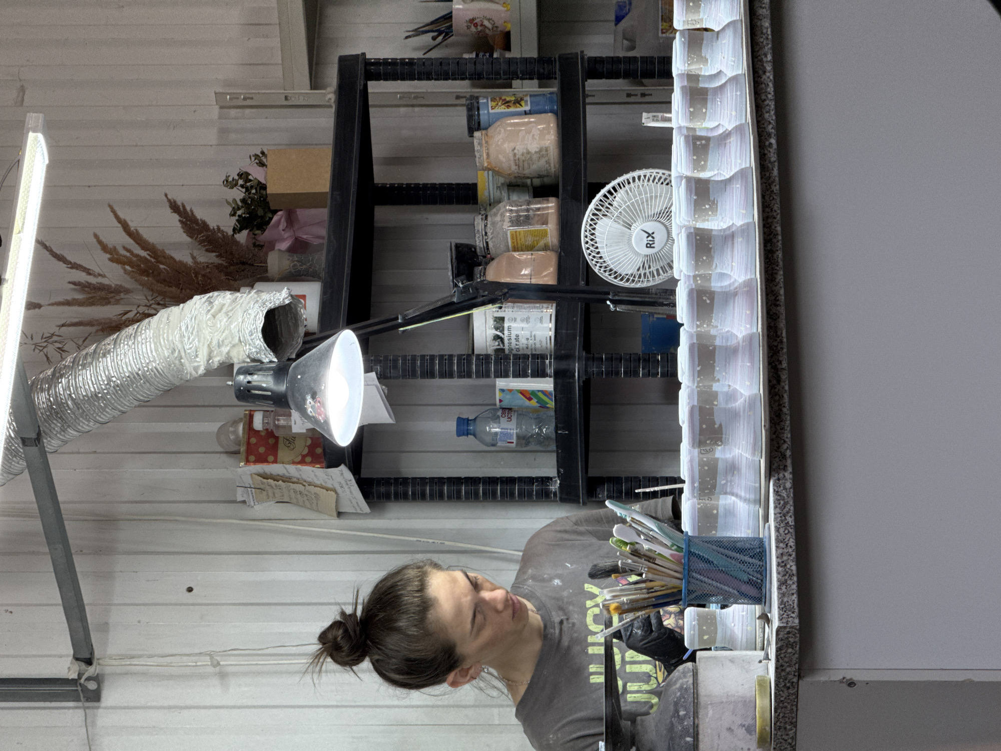 A person with brown hair in a bun is standing in a room that appears to be a workshop or studio, as suggested by the abundance of art supplies and shelving units filled with various objects. The room has a white, possibly corrugated metal wall, typical of industrial buildings.
The subject is wearing a dark grey t-shirt and is positioned to the left, facing towards the right of the frame. A light fixture with a silver, corrugated flexible duct is directed at the work area. Above the person, there's a black metal shelving unit filled with jars, containers, and other items. A small white fan is visible on the shelf.
On the right side of the image, there's a vertical stack of what appears to be molds or castings along with a metal container overflowing with brushes. A large light grey wall or partition is on the right edge of the image.
Based on the prompt, the location is in Chaikovsky, Russia.