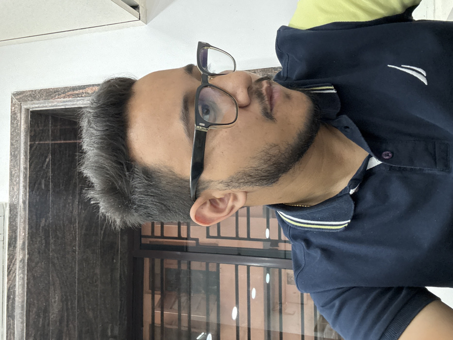 The image presents a head-and-shoulders portrait of a single male individual, likely a young adult, situated indoors in Kolkata, India. The subject has dark, textured hair styled with volume, fair to medium skin tone, a neatly trimmed beard, and a mustache. He wears clear-framed eyeglasses with dark upper rims. His attire is a dark blue polo shirt with a three-button placket and a white and yellow striped collar. A white sailboat logo is embroidered on the left chest, and a slender gold chain is visible around his neck. The individual is positioned with his body facing slightly forward and his head turned to his right, looking off-camera with a neutral expression. The background features a reflective, dark surface on the left, possibly a window or framed glass, showing indistinct horizontal and vertical lines and light reflections. Above this, a dark, veined stone surface is present. To the right, a plain white wall or cabinet unit is visible.