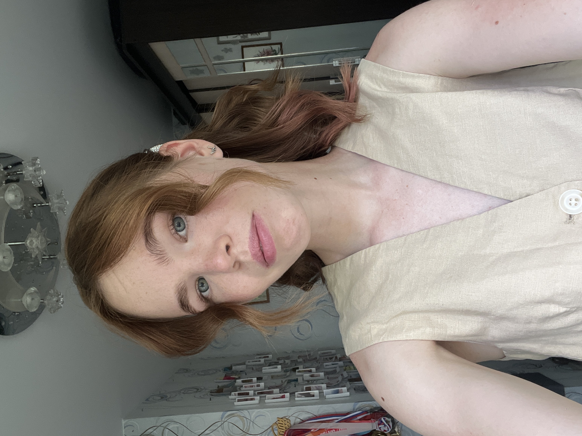 In a selfie taken from a slightly high angle, a young woman with fair skin, freckles, and wavy reddish-blonde hair is centered. She has light blue eyes and is wearing a beige sleeveless vest with a V-neckline and a single white button visible at the bottom. Her hair is pulled back loosely, with some strands framing her face, and the tips have a pinkish hue. She is wearing a small silver earring in her left ear. The background suggests an indoor setting, possibly a room in Chaikovsky, Russia. To her left, a wall is adorned with a collage of small framed photos and a cluster of medals. Above her, a chandelier with multiple glass elements hangs from the ceiling. To her right, a tall, dark wardrobe with mirrored panels is visible, reflecting some of the room's interior. The lighting appears natural, coming from the front, illuminating her face and upper body.