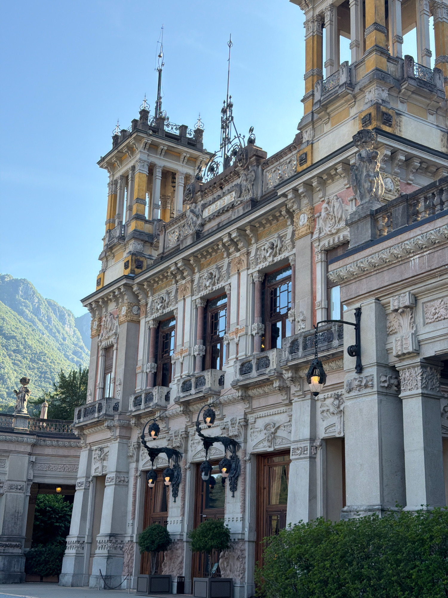 The image shows the exterior of the historic San Pellegrino Casino in San Pellegrino Terme, Italy. The grand Art Nouveau building features ornate architectural details, including columns, balconies, and decorative sculptures. The facade is painted in shades of yellow and white, with intricate stonework and large windows. In the background, lush green mountains rise against a clear blue sky. The foreground is occupied by manicured bushes and trees, with elaborate outdoor lamps adorning the building's facade. The overall scene depicts a sunny day with the historic casino as the prominent subject.