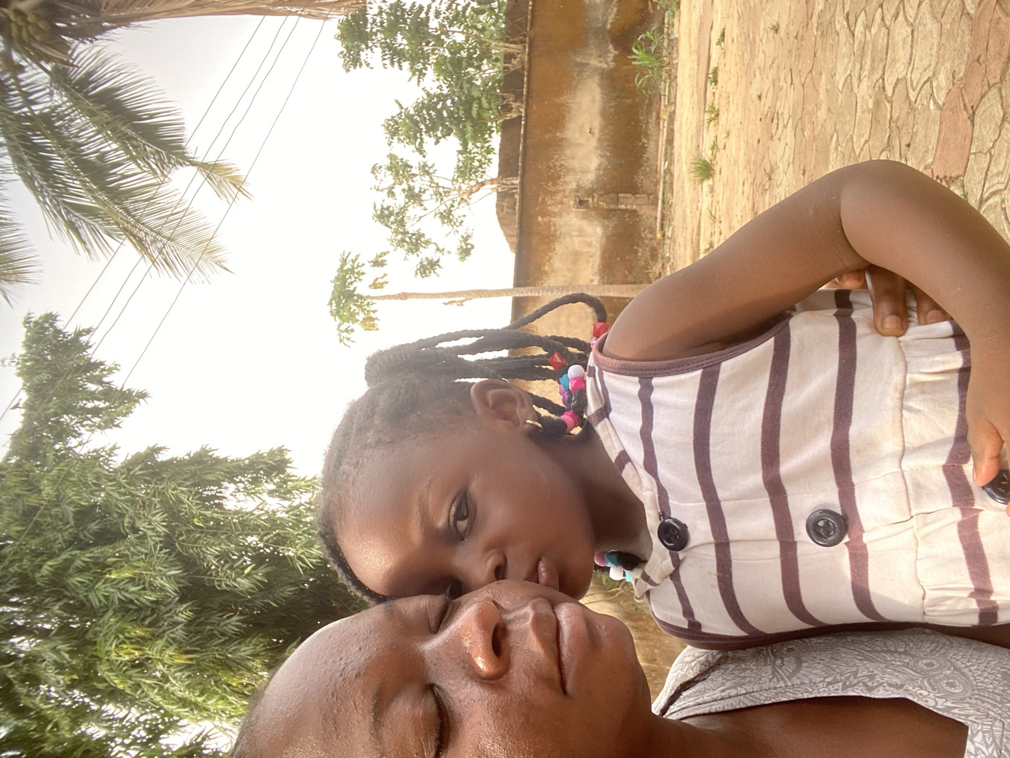 A young girl with beaded braids rests her face on an adult, likely a mother, against a backdrop of a sunny outdoor scene. The girl wears a striped white and brown dress with buttons. The background includes palm trees, a wall, and other vegetation, suggesting a location in Maigizo, Nigeria.