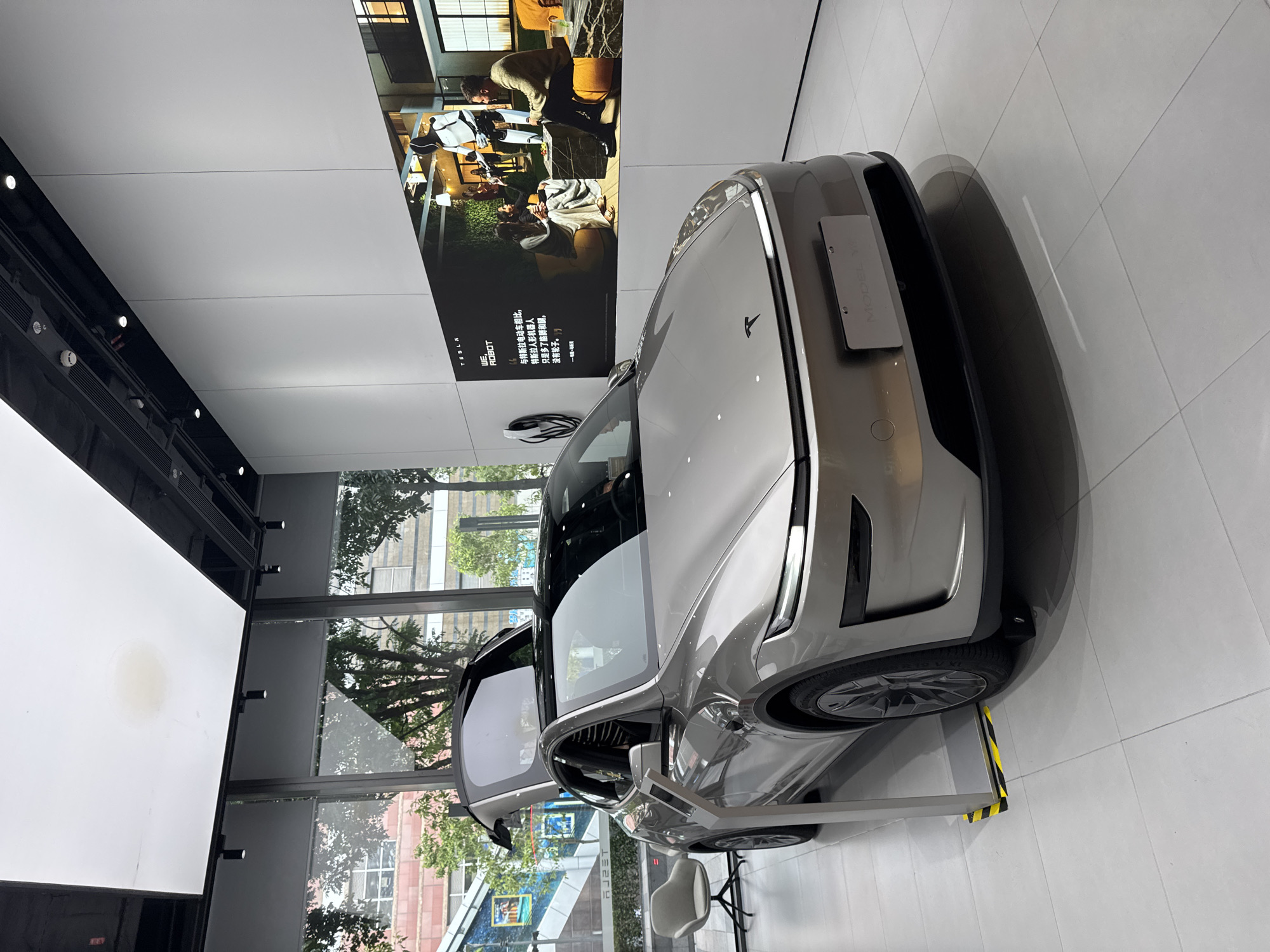 The image shows a gray Tesla Model Y car displayed in a showroom in Tianhe District, China. The car is parked on a white tile floor, partially on top of a yellow and black striped block. One of the car doors is open. In the background, there is a large poster displaying a picture of people at a table with a robot; next to it is the Tesla logo and some Chinese text. The poster reads "TESLA ROBOT," "与特斯拉电动车相比,特斯拉人形机器人只是多了胳膊和腿, 没有轮子,一号", and "MODEL Y". Large windows reveal an exterior scene with trees and buildings. A chair sits near the car.