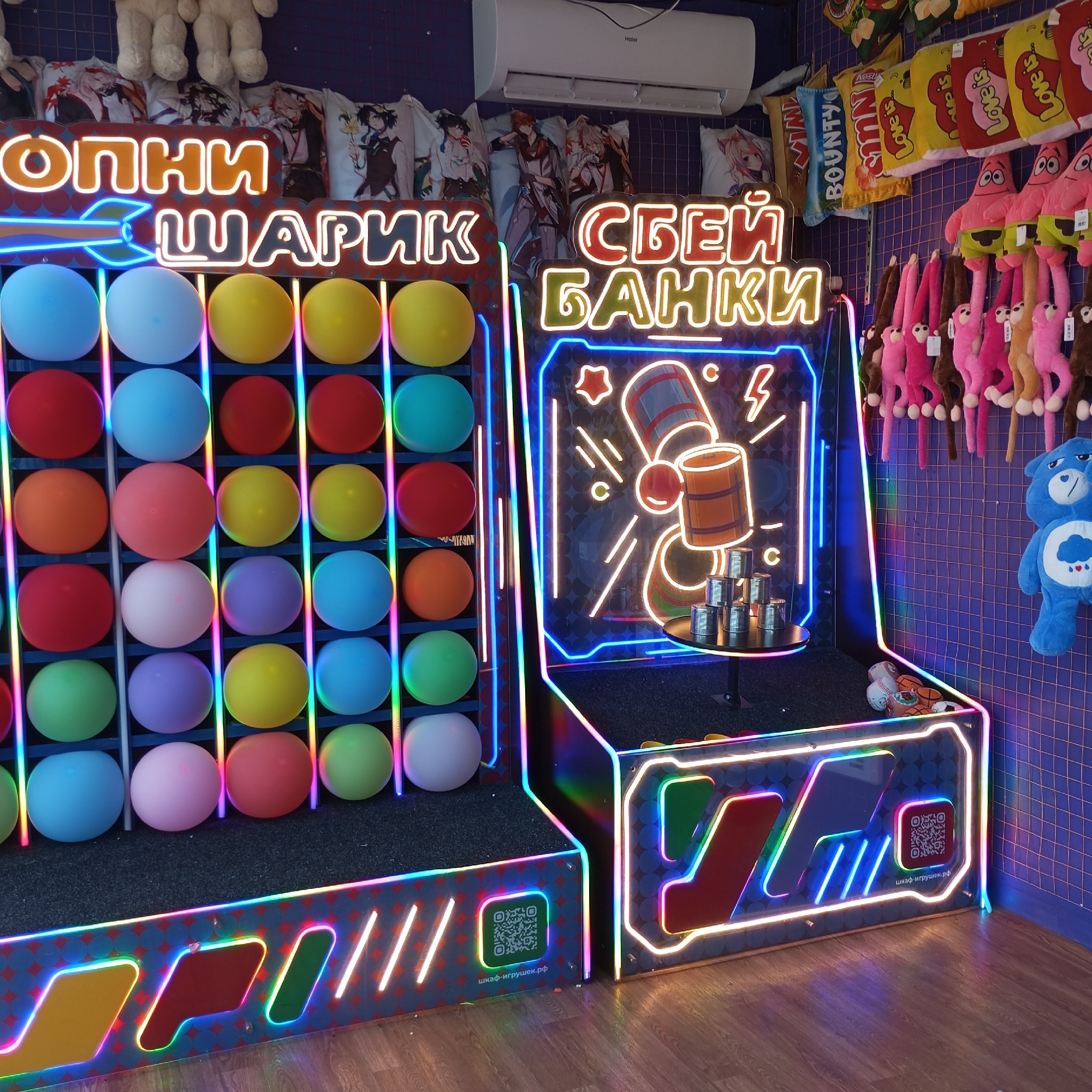 This image captures an indoor arcade or game area in Pervomaysky, Russia, featuring two neon-illuminated game stations and various prizes displayed on a dark blue grid-patterned wall.
On the left, a game station displays "ЛОПНИ ШАРИК" (Pop the Ball) in orange neon Cyrillic letters. Below the text, a grid structure holds numerous colorful, spherical objects (balls or balloons) arranged in rows, illuminated by vertical neon light strips. Colors include light blue, white, yellow, red, orange, pink, purple, and green. The base of this station features multi-colored neon outlines over geometric patterns and a QR code with "шкаф-игрушек.рф" printed below it.
On the right, a second game station displays "СБЕЙ БАНКИ" (Knock Down Cans) in red and green neon Cyrillic text. An internal neon graphic depicts a cannon aiming at stacked barrels. A small circular black table below the graphic holds a pyramid of six silver-colored cans. The base of this station also presents multi-colored neon outlines and geometric patterns. A baseball and a basketball are visible on the floor near its base.
The wall behind the game stations is adorned with prizes, including a row of anime-themed body pillows (featuring characters such as Venti and Childe from Genshin Impact), assorted pillows designed to resemble snack packaging (e.g., Bounty, Nestle, Love is...), multiple pink Patrick Star plush toys, several long-limbed monkey plush toys in pink, brown, and peach, and a large blue Care Bear plush toy (Grumpy Bear). A white Haier air conditioning unit is mounted on the wall above the anime pillows.
The floor is constructed of light brown wood, exhibiting reflections from the neon lighting. No individuals are present in the scene.