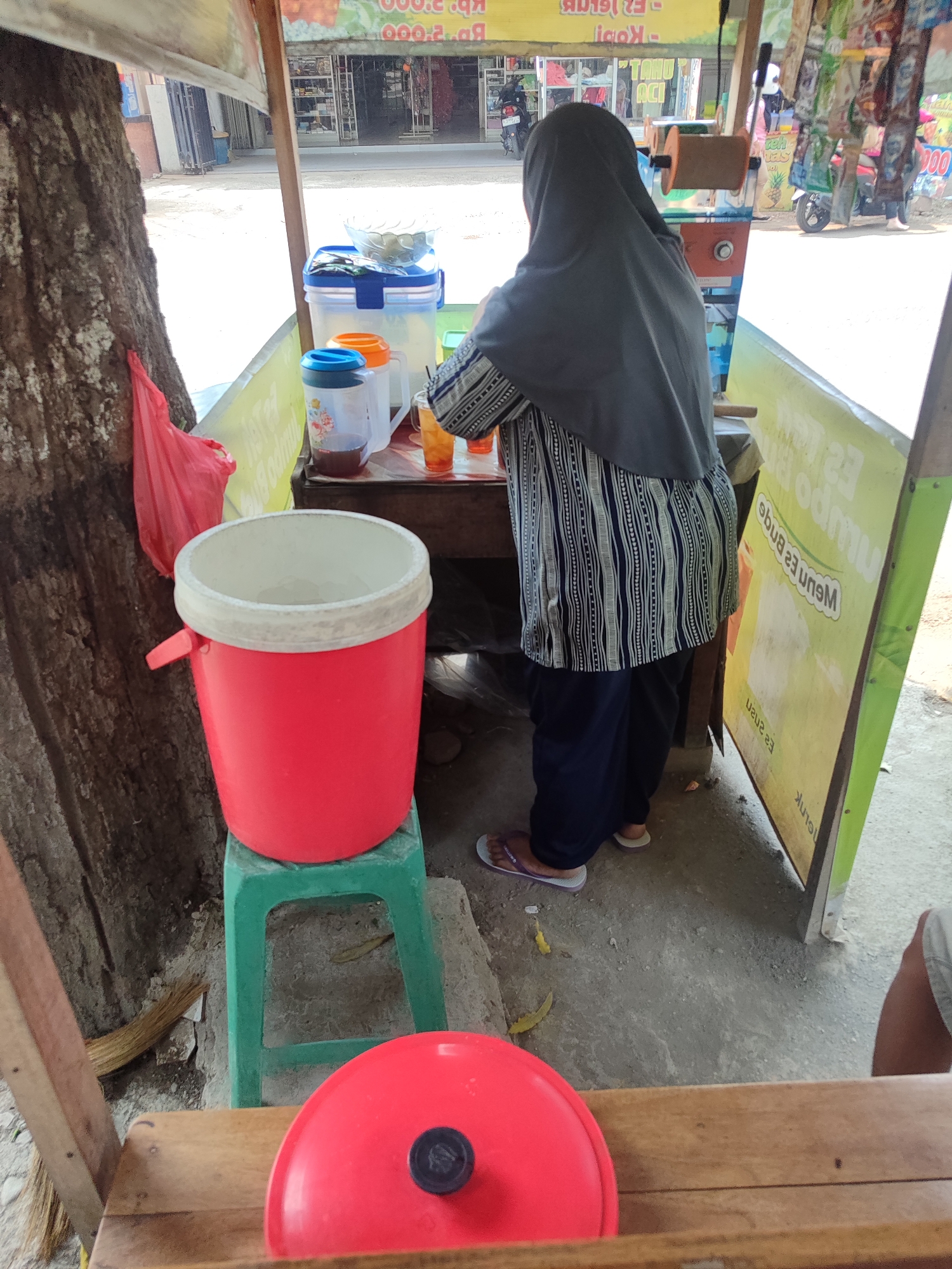 A woman wearing a dark grey hijab and a patterned blue and white tunic is standing behind a mobile food stall. The stall is made of wood and has a yellow banner with text in Indonesian. In front of the woman is a red cooler bucket with a white rim, placed on a green plastic stool. There are also two clear plastic pitchers with lids, one containing a dark liquid and the other with an orange liquid. Several plastic cups are on the counter. To the left of the stall, a large tree trunk is visible, with a red plastic bag hanging from it. The ground is a dirt or gravel surface. In the background, there are buildings and motorcycles, suggesting an outdoor urban or semi-urban environment, likely a street or market area in Kagok, Indonesia.