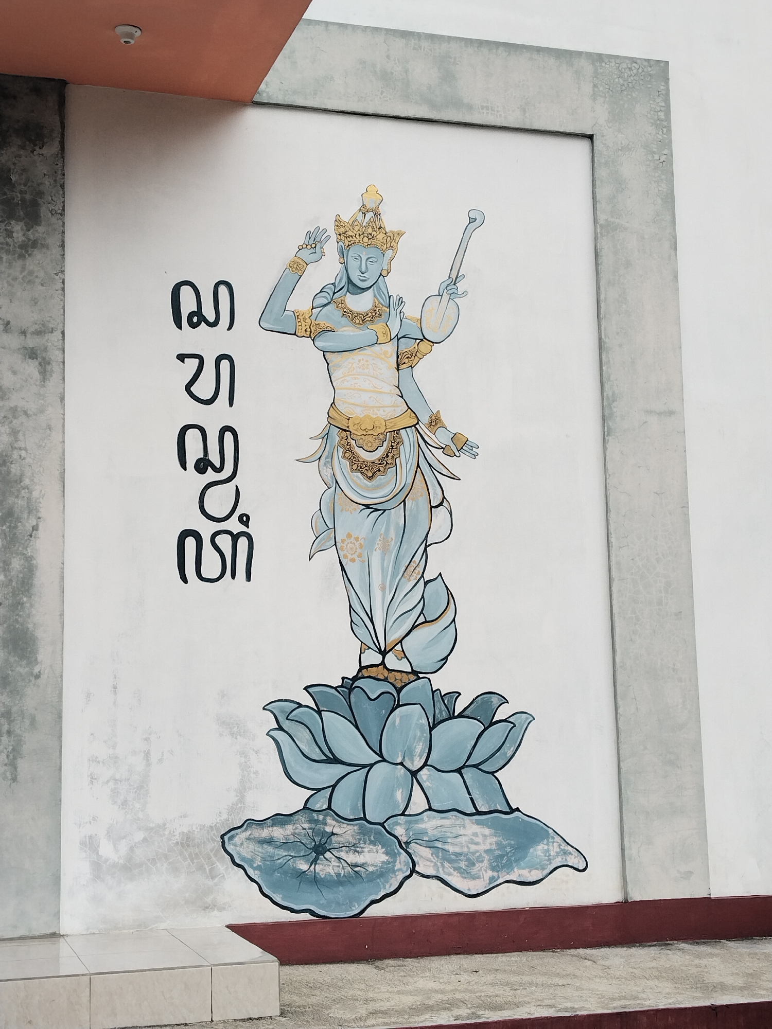 A vertical, eye-level shot captures a mural of a four-armed figure mounted on a white wall flanked by grey architectural elements. The figure, rendered in a bluish hue with gold accents, stands atop a large lotus flower. The figure appears to be a representation of a deity, possibly Saraswati, given the musical instrument and hand gestures. To the left of the figure, a series of vertical symbols are rendered in black. The wall is framed by what seems to be a concrete or stone structure, indicating the mural is part of a building's exterior. The floor is visible at the bottom, showing beige tiles and a grey concrete area.