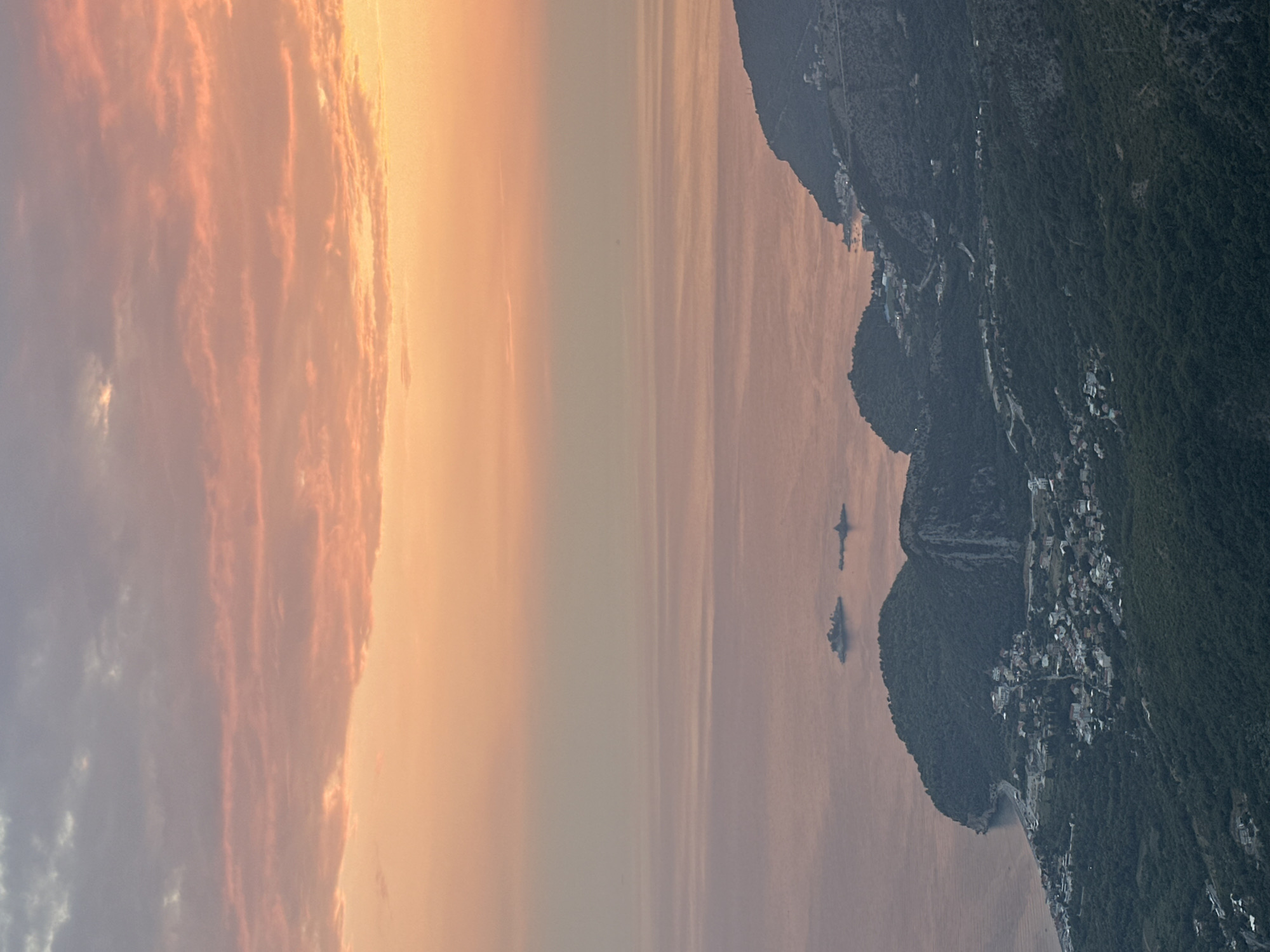 This is a vertical, high-angle aerial shot of the coast of Gluhi Do, Montenegro at sunset. The sky is a gradient of pale orange and pink with thin, wispy clouds. The sea is a calm, reflective surface, with a hazy horizon line. In the distance, two small, rocky islands are visible in the water. Closer to the shore, a lush, green coastline curves inwards. A small town is nestled along the coast, with white buildings and red roofs visible amongst the trees. The town appears to be built on a series of hills that slope down towards the sea. The surrounding landscape is densely forested with dark green trees, creating a stark contrast with the bright sky and calm water. The overall impression is one of a peaceful, remote coastal community bathed in the soft light of sunset.