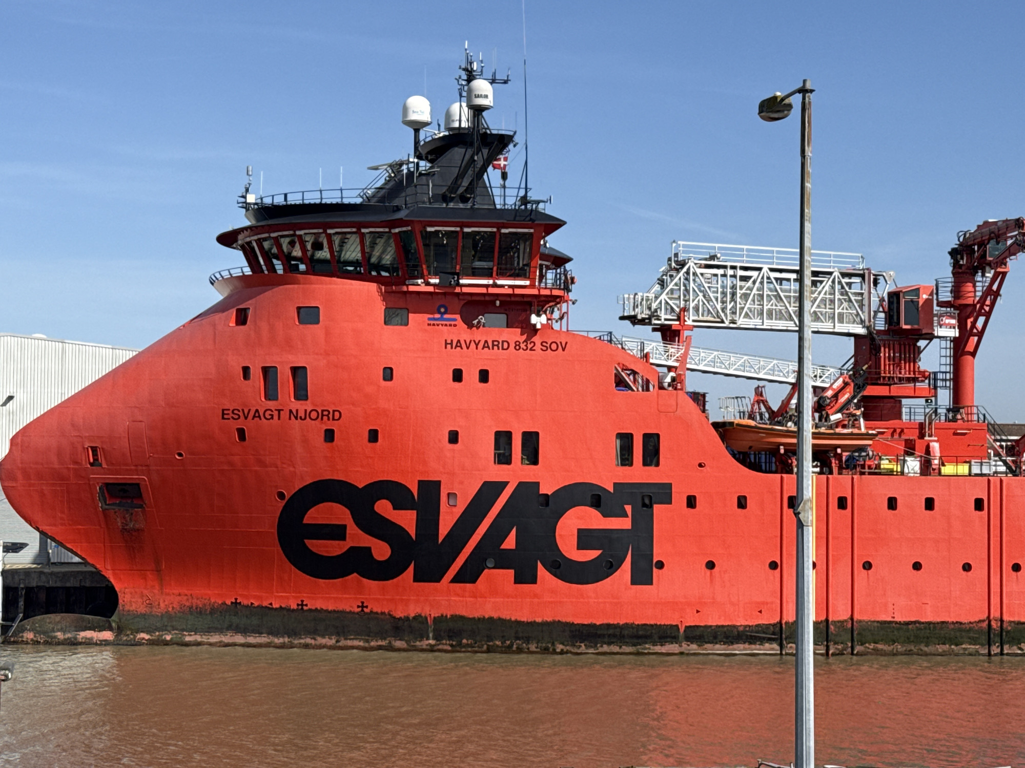 The image shows a large red ship, the "ESVAGT NJORD HAVYARD 832 SOV", docked in Gorleston-on-Sea, United Kingdom. The ship has a prominent black "ESVAGT" logo on its hull. Above the ship, there is a gangway structure in white and red. A gray lamppost stands in the foreground, partially obscuring the view. The water in the foreground appears brown. The sky is clear and blue. The architectural elements of nearby buildings can be seen, indicating a port or industrial area.