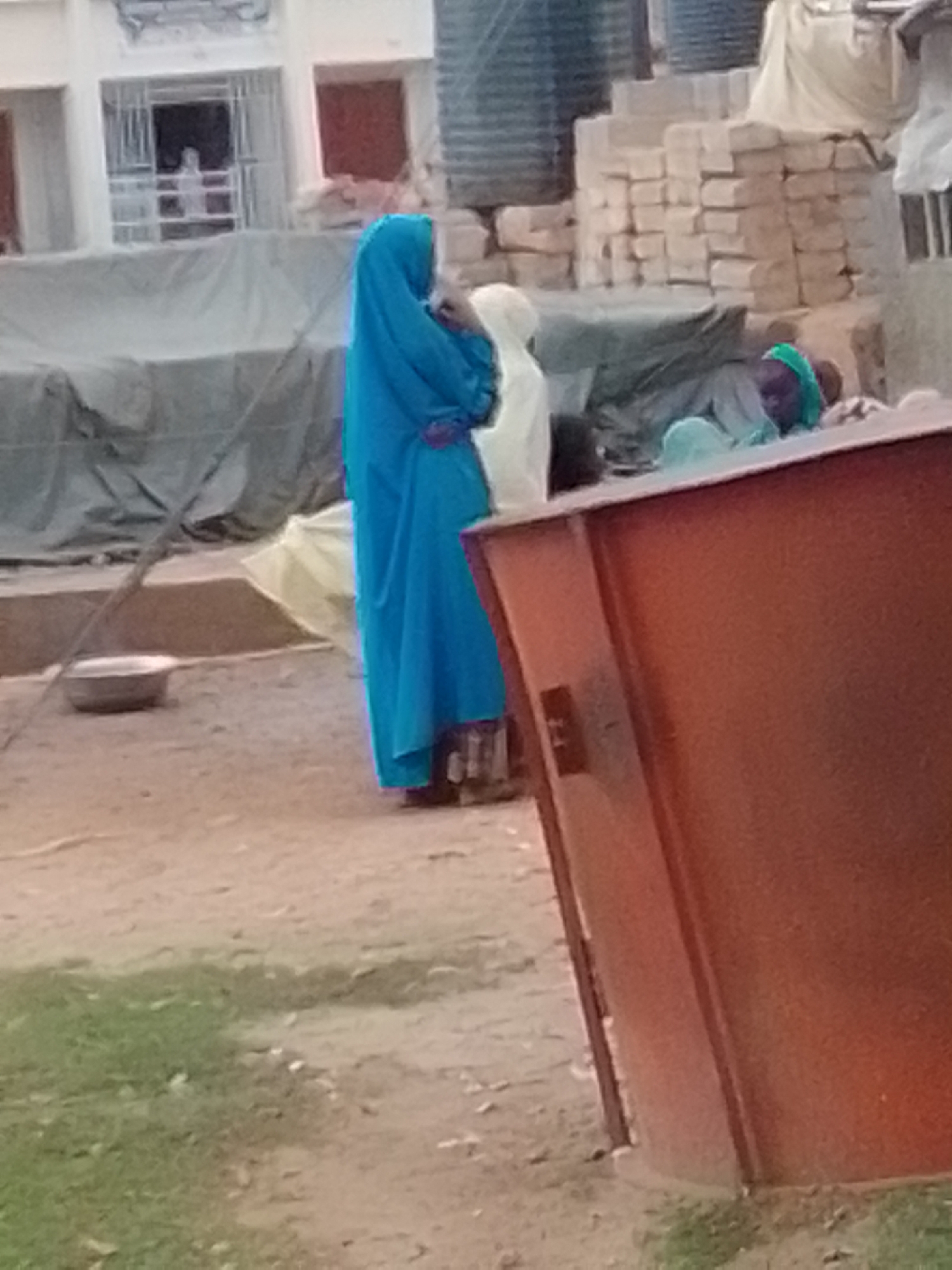 In Pantame, Nigeria, a person wearing a bright blue hijab and a long blue garment stands on a dirt path. To their right, another person is partially visible, wearing a green headscarf and a light-colored garment. In the background, there are stacks of bricks, large cylindrical tanks, and a tarp-covered structure. A small metal bowl sits on the ground in front of the person in blue. The scene appears to be outdoors, possibly in a market or construction area, with dry earth and sparse grass.