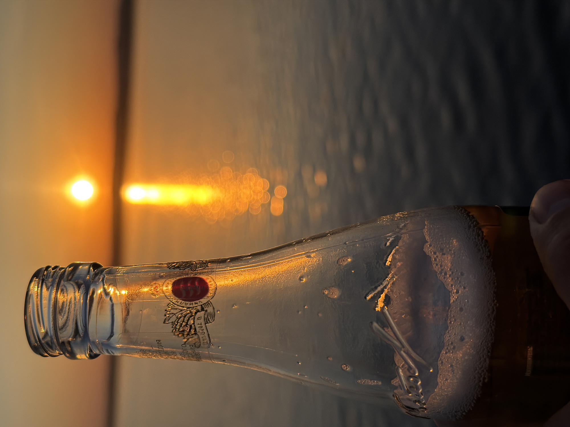 The image shows a partially consumed bottle of beer held up against a sunset over water. The bottle is clear glass with condensation and visible bubbles of beer foam inside. The bottle's label includes some writing in Russian and what appears to be a red emblem. The background features a body of water with the sun setting or rising in the distance, reflecting a bright orange streak across the water. The image suggests a peaceful moment. The city of Chaikovsky, Russia, is not evident in the image.