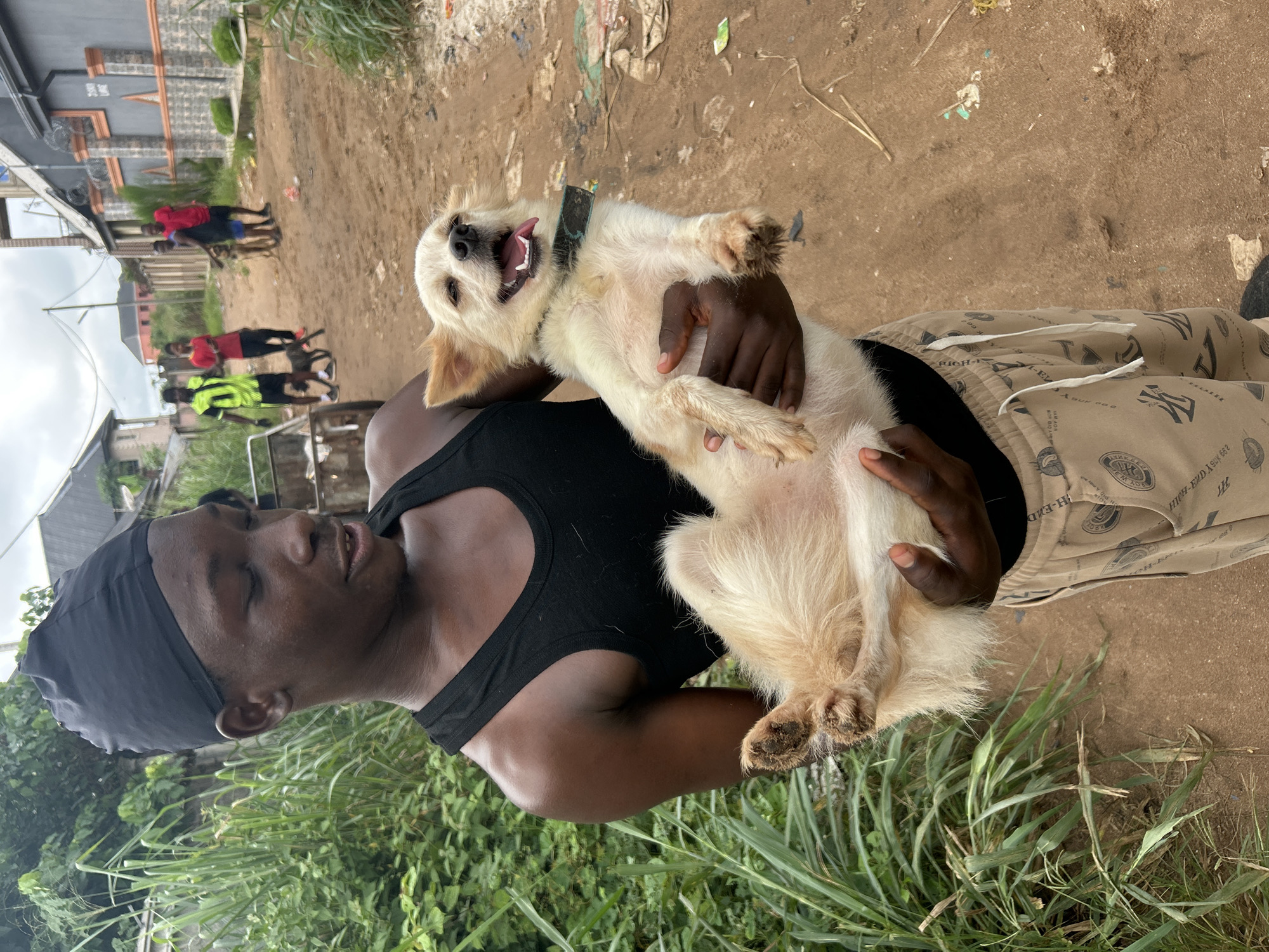 In the city of Ihiagwa, Nigeria, a young Black male wearing a black durag and a black tank top holds a small, light-colored dog. The dog, with its mouth open and tongue out, appears to be panting. The man is wearing light-colored sweatpants with a repeating pattern of black graphics and text. In the background, several other individuals are visible, along with a metal cart. The environment consists of a dirt path lined with tall green grass and buildings. The sky is overcast.
