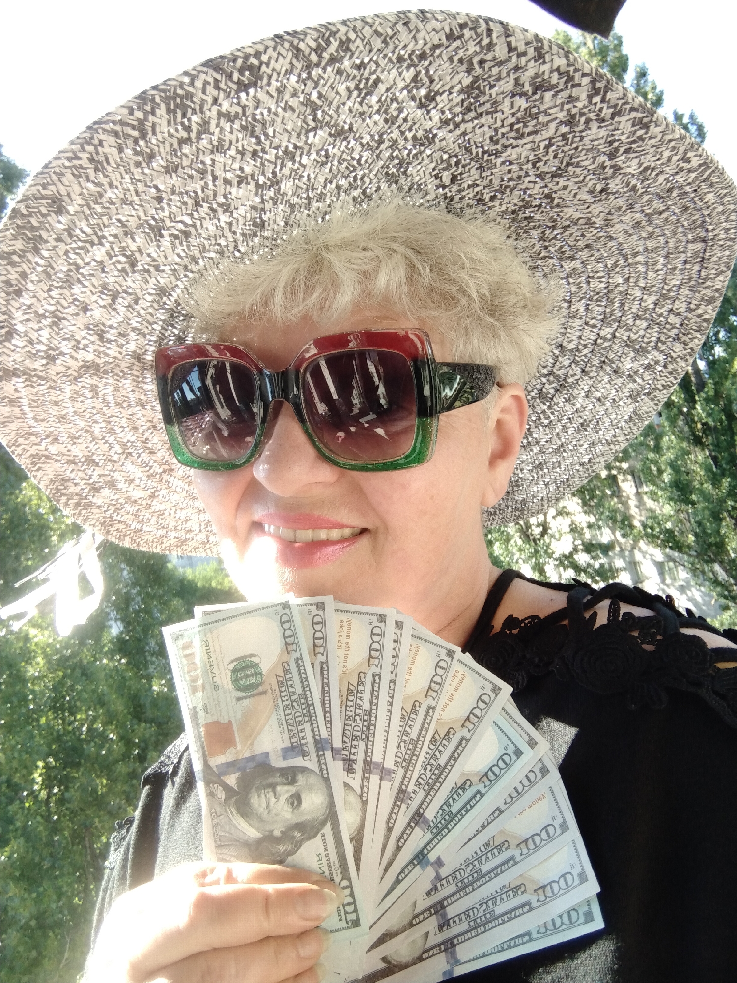 A medium shot captures an older woman wearing a wide-brimmed straw hat and oversized sunglasses with red and green frames. She is holding a fan of US $100 bills. She is smiling and looking towards the camera. The background shows green trees and foliage, suggesting an outdoor setting. The scene is not identifiable as being in Horishni Plavni, Ukraine.