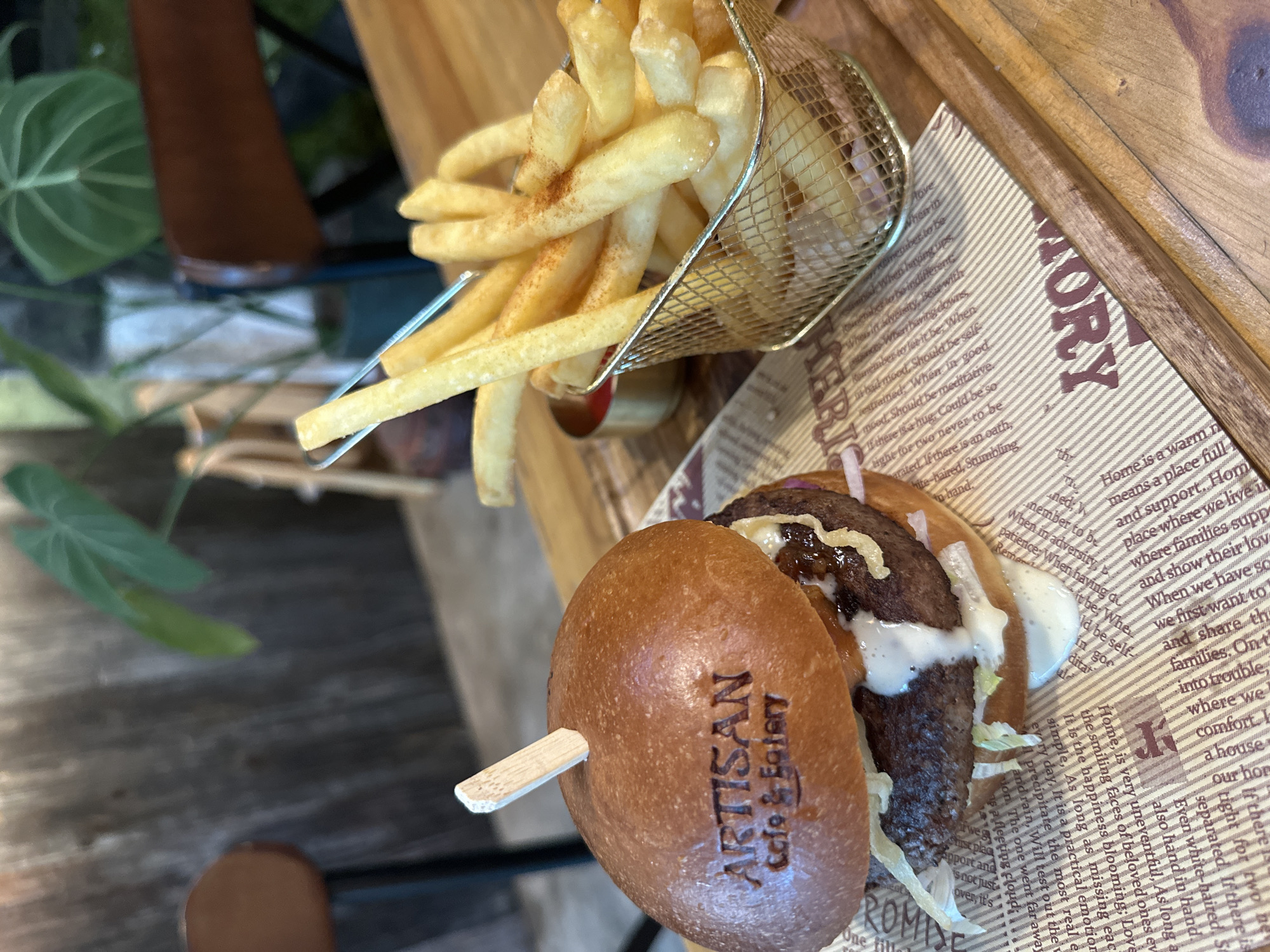 A close-up, top-down view captures a burger and fries served on a wooden tray lined with patterned paper. The main subjects are a savory burger with a thick patty, white sauce, and shredded lettuce, accompanied by a side of golden-brown fries in a miniature metal frying basket. A wooden skewer pierces the burger, and its top bun is branded with "ARTISAN Cafe & Eatery". The fries appear lightly seasoned.
The paper lining the wooden tray features various texts, including prominent words like "MORY", "HOME", "HERIG", and "PROMISE". Numerous sentences are legible, discussing themes such as the meaning of "Home" as a place of warmth, support, and love for families. Other texts offer advice on handling life's ups and downs, self-restraint, meditation, and the value of hugs and family bonds.
In the blurred background, hints of green foliage from a plant are visible, suggesting an outdoor or semi-outdoor dining setting. The natural lighting indicates it is daytime. The meal is presented in a casual yet inviting manner, likely at a cafe or eatery in Nha Trang Ward, Vietnam, as indicated by the context.