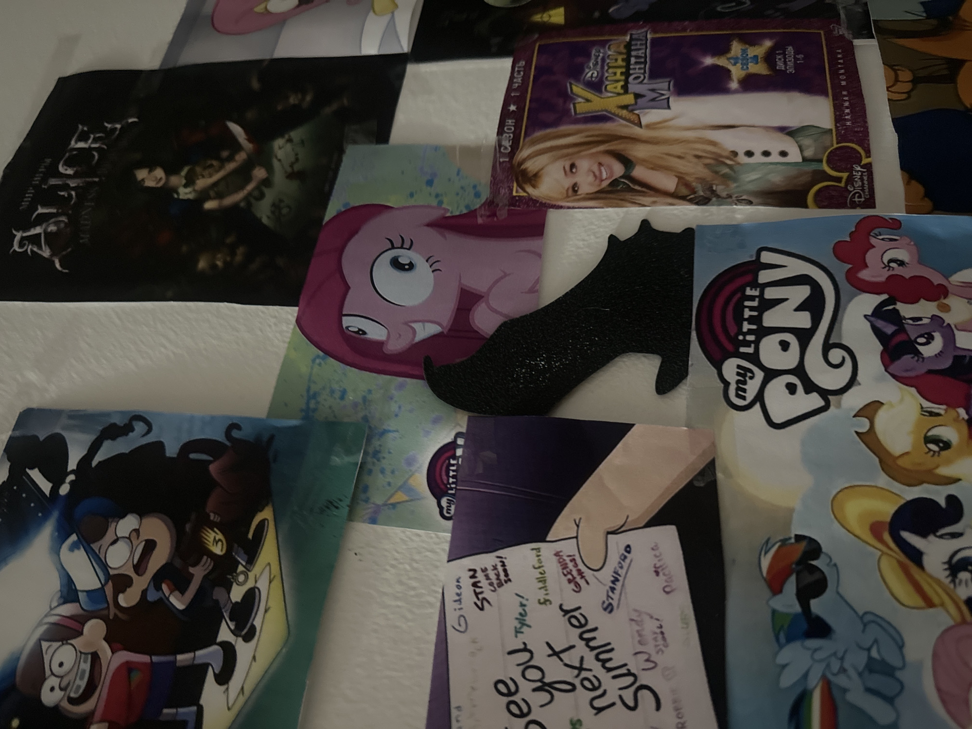 This flat lay image displays a collection of fan merchandise and media, predominantly featuring elements from popular animated series and video games.
In the upper left, a dark-themed poster or print for "Alice: Madness Returns" is visible, showing the titular character, Alice, holding a knife. Below and to the right of this, a vibrant print of Pinkie Pie from "My Little Pony" with a wide-eyed, surprised expression can be seen.
Above Pinkie Pie, a DVD cover for "Hannah Montana" is prominently placed. It features Miley Cyrus as Hannah Montana, smiling. Visible text on the DVD cover includes "Disney CHANNEL," "HANNAH MONTANA," and Russian text indicating "1 СЕЗОН 1 ЧАСТЬ" (1st Season, 1st Part) and "ДИСК 1 ЭПИЗОДЫ 1-6" (Disc 1, Episodes 1-6).
To the right, a large print or poster for "My Little Pony" showcases several main characters, including Rainbow Dash, Twilight Sparkle, and Applejack, along with the "my LITTLE PONY" logo.
In the bottom left, a poster featuring Dipper and Mabel from "Gravity Falls" is visible, depicting them in an animated, action-oriented pose. Overlapping this poster and extending towards the center is a piece of white paper with handwritten messages. The text, in various handwriting styles, mentions multiple characters from "Gravity Falls," such as "Gideon," "STAN COME BACK SOON!," "Tyler!," "Fiddleford," "GLENDA," "STANFORD," "Wendy STAY cool!," and "Pacifica." Phrases like "see you next Summer" are also legible.
A black, glittery cutout in the shape of a high-heeled shoe is placed centrally, overlapping the Pinkie Pie print and extending towards the "My Little Pony" poster. The items are arranged on a light, possibly white, textured surface, suggesting a display or collection.