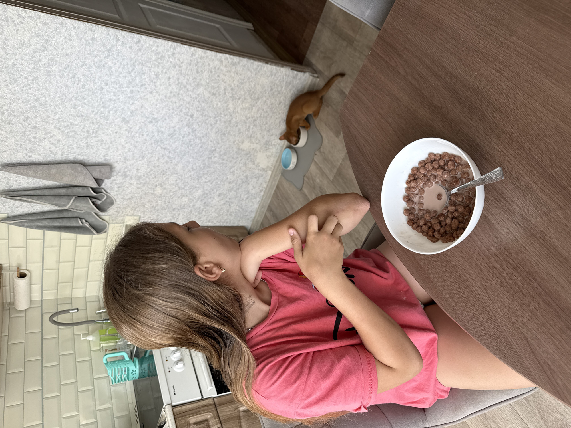 A young girl with long brown hair is seated at a wooden table in a kitchen, looking to her right. She is wearing a pink t-shirt and appears to be contemplating something, as her hand is resting on her cheek. On the table in front of her is a white bowl filled with chocolate cereal and milk, with a spoon inside. In the background, a small, reddish-brown cat is seen eating from a bowl on the floor. The kitchen has white tiled walls and a patterned wallpapered section. There is a stove and sink visible in the background. The scene is indoors, likely in a residential kitchen. The image does not provide any specific details that would allow for an accurate location in the city of Stary Oskol, Russia.