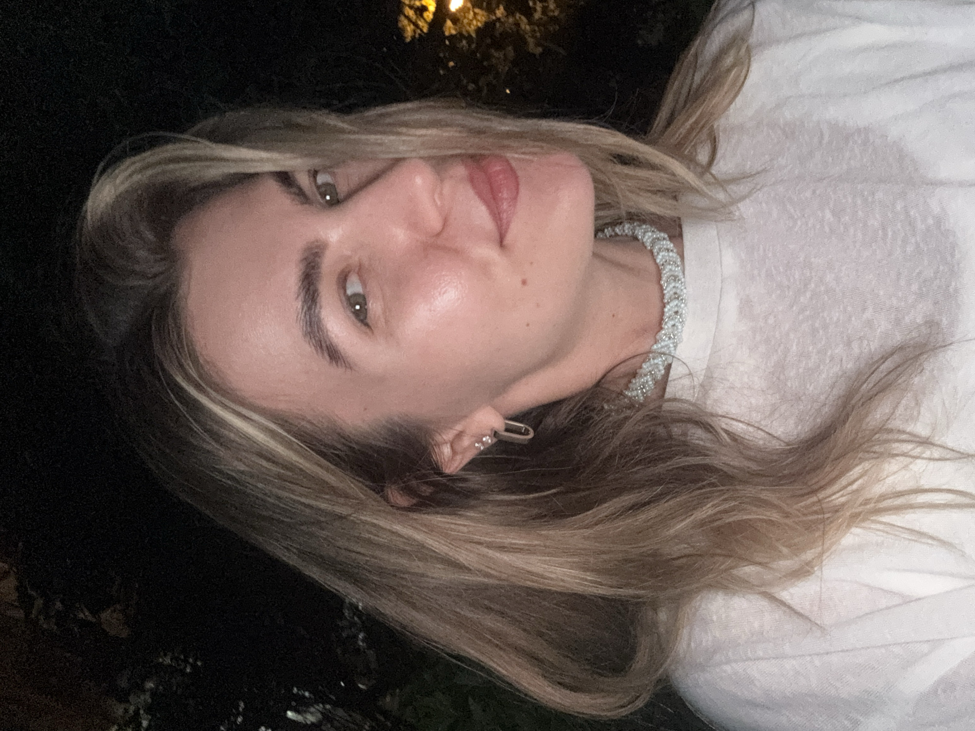 A close-up, low-light portrait of a woman with blonde hair is shown from the chest up. She is wearing a white t-shirt and a beaded necklace. She is looking to the right and smiling. Her hair is long and wavy, and she is wearing earrings. The background is dark, with some foliage and lights visible on the right side. The image appears to be taken outdoors at night.