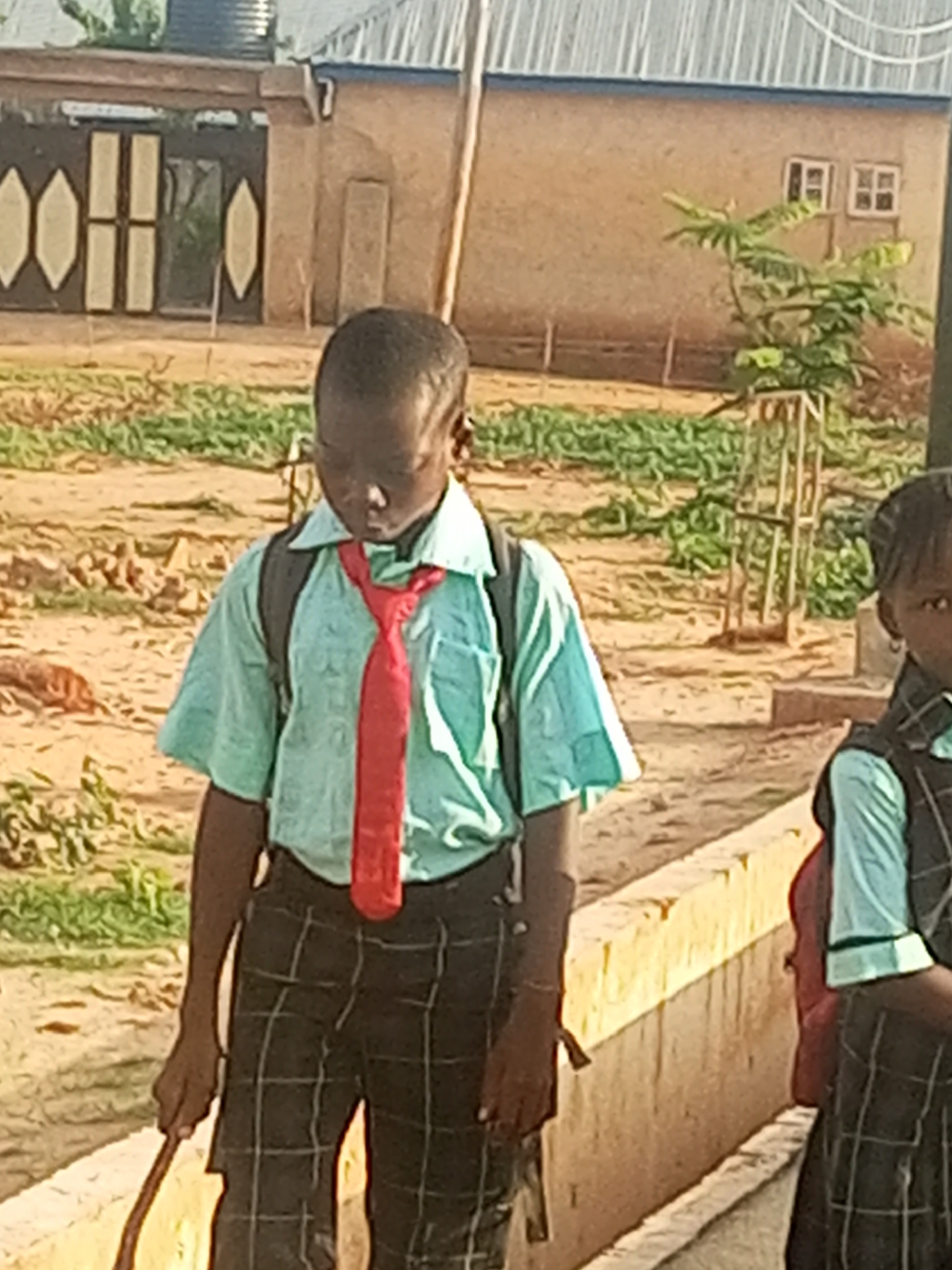 A young Black child wearing a light blue collared shirt, a red tie, and plaid pants stands in front of a building with a corrugated metal roof. The child is looking down and to their left, with a black backpack on. Another child, also wearing a plaid uniform and a light blue shirt, is partially visible to the right of the frame. In the background, a tan building with two small windows is visible, along with some green foliage and a fence. The ground appears to be dirt and gravel.