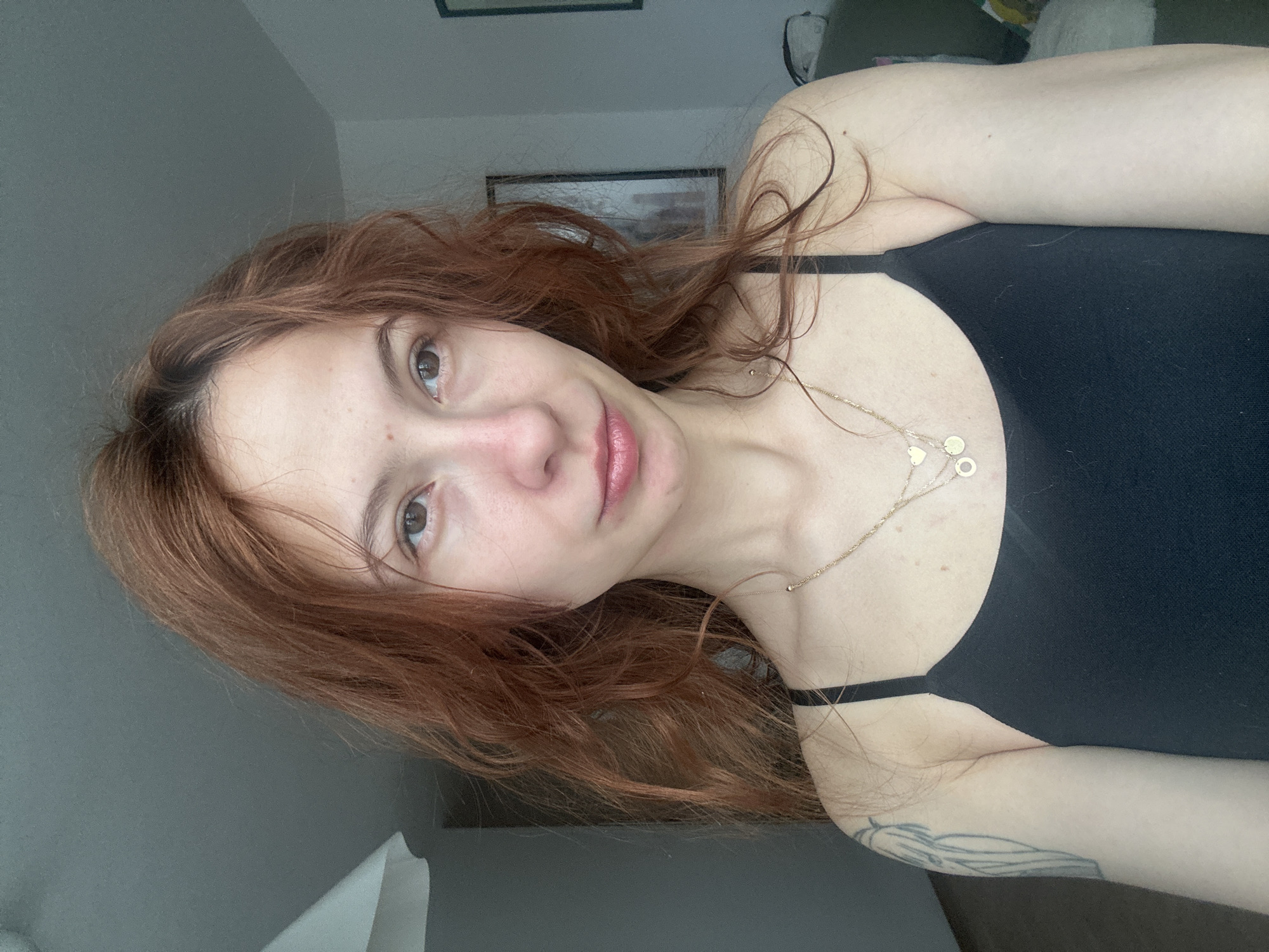 A woman with red hair is wearing a black tank top and a gold necklace with circular pendants. She has fair skin and brown eyes. The background shows a wall, a picture frame, and a glimpse of furniture, possibly indicating an indoor setting. There is no indication of the image being in Novi Sad, Serbia.