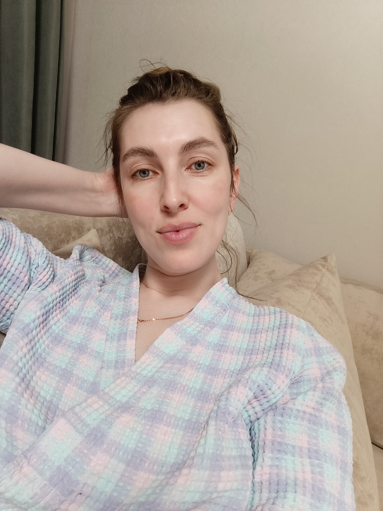 A woman is reclining on a beige couch, wearing a pastel-colored, checkered robe. Her hair is pulled back into a bun, and she is wearing a thin gold necklace. Her left hand is behind her head. The background includes a beige wall and a green curtain. The location is Chaikovsky, Russia.