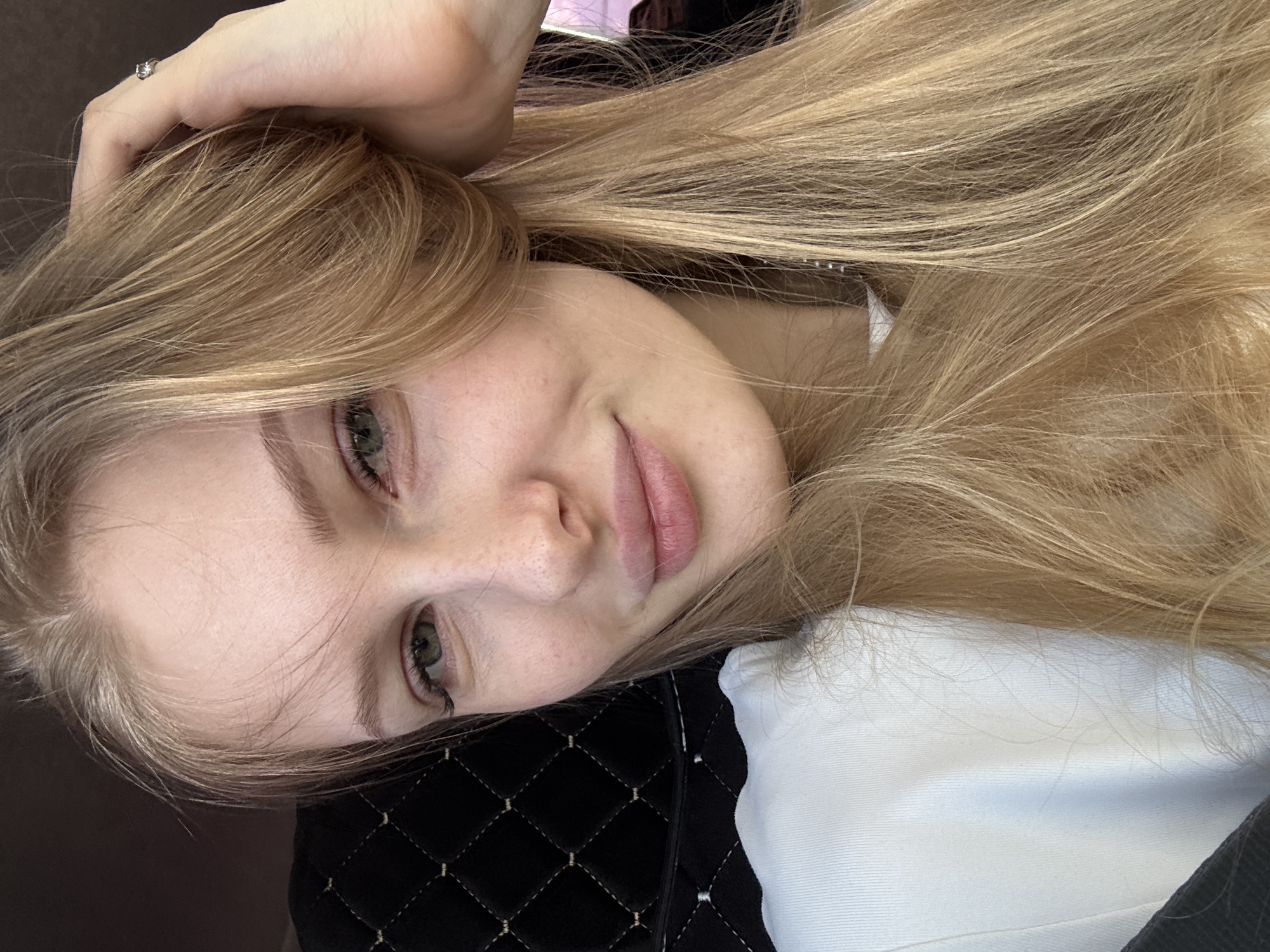An angled, close-up portrait features a young woman with long, straight, light blonde hair. Her head is tilted, resting on a dark, quilted surface with a diamond pattern and lighter stitching. She looks directly at the camera with light-colored eyes, defined eyelashes, and a gentle, relaxed smile on her naturally pink lips. Her fair skin shows a scattering of freckles across her nose and upper cheeks.
Her left hand is positioned near her head, with fingers lightly touching her hair. A silver-colored ring with a small, clear gemstone is visible on her left ring finger. She is wearing a white top, a portion of which is visible on the lower right of the frame. The lighting is soft and even, suggesting an indoor setting during the daytime. The overall mood is calm and candid. No text is visible in the image. The location context for this media file is Perm, Russia.