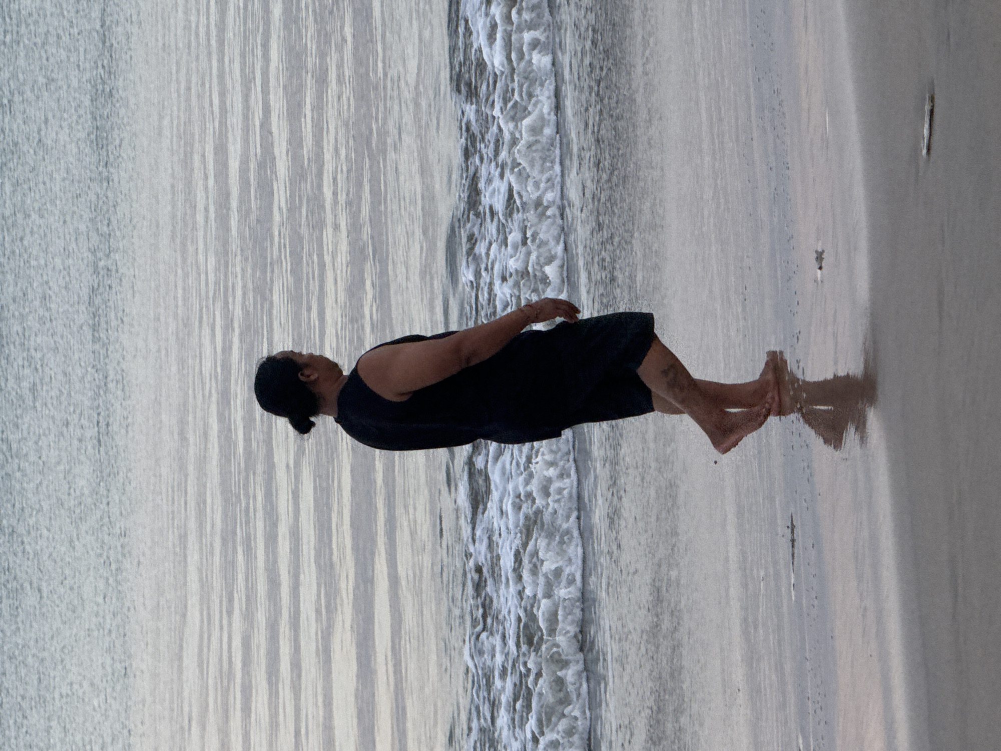 A person wearing a black sleeveless shirt and dark shorts walks along the shoreline of Kuta Beach, Indonesia. The person's hair is pulled back in a bun. The sea is calm with gentle waves breaking on the sand. The sand is wet and reflects the sky. The scene is tranquil and appears to be during either dawn or dusk.