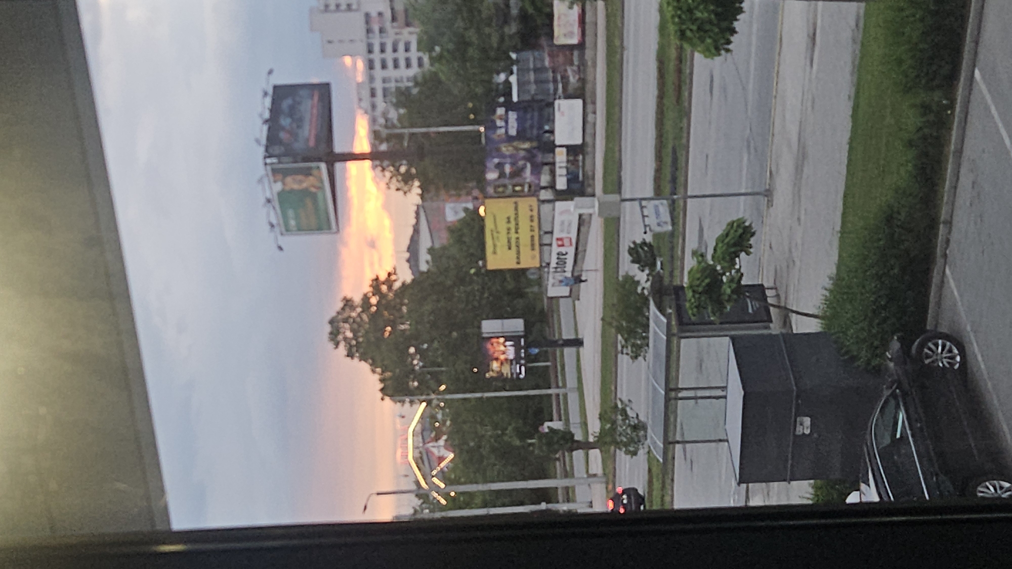 The image depicts an urban view from an elevated perspective, likely a window. A street runs horizontally through the frame, bordered by greenery on one side and buildings with various advertisements on the other. The sky shows a gradient of colors suggesting either a sunrise or sunset. Several billboards and advertisements are visible along the street. A black car is parked on the right side of the frame, and a bus stop or shelter is nearby. In the background, a building appears, which is likely the "Hotel St.Petersburg" in the city of Plovdiv.