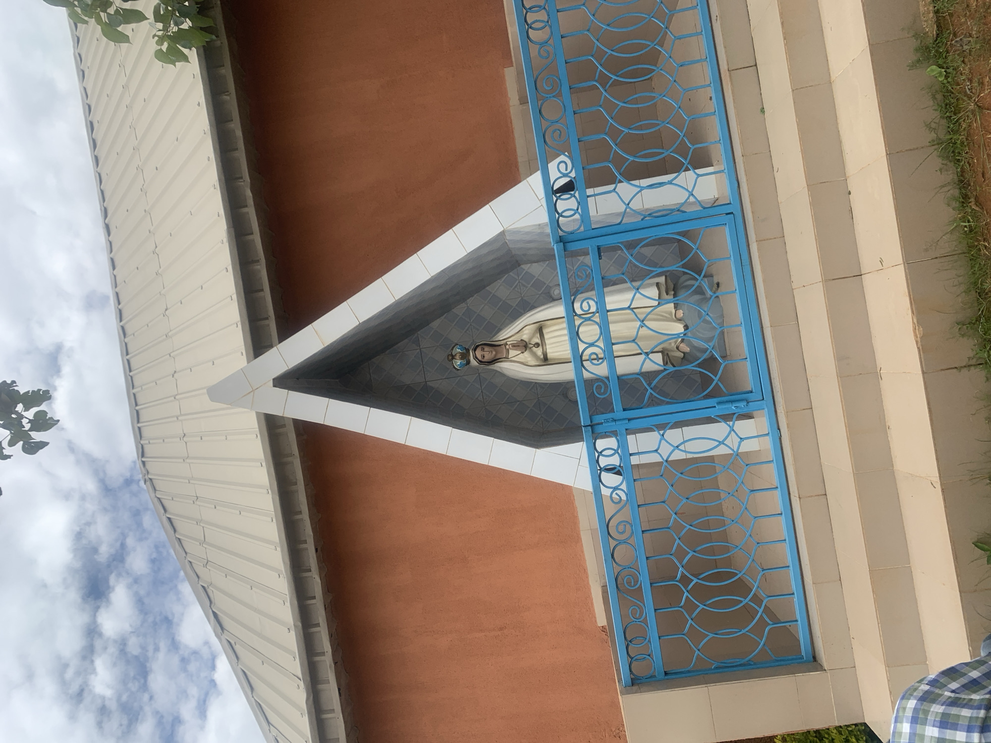 The image shows a statue of the Virgin Mary with a crown, dressed in white robes, standing in a niche. The niche is triangular in shape with white trim and a patterned background. In front of the niche is a blue metal fence with a decorative pattern. The statue is on a raised platform with steps leading up to it. Behind the niche and statue is an orange building with a beige metal roof. The sky is cloudy. The scene appears to be outdoors, possibly in a public space or religious site. The location is identified as Duk, Nigeria.