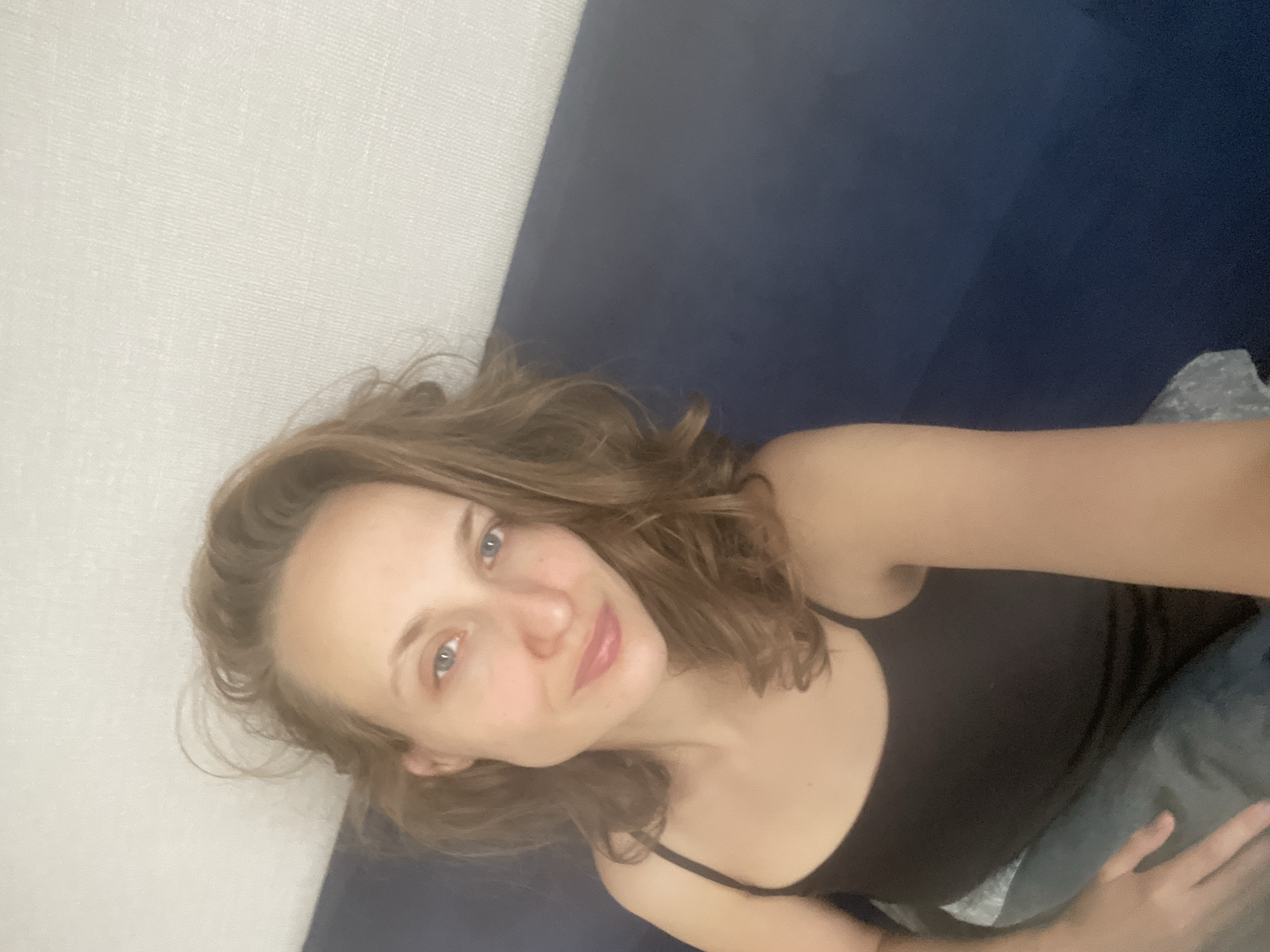 A young woman with light brown, wavy hair is seen in a close-up selfie, looking directly at the camera. She is wearing a black tank top and appears to be indoors, possibly on a couch or bed, indicated by the blue upholstery behind her and a patterned textile beneath her. The background consists of a textured white wall. The lighting is soft, and her expression is neutral. No specific location in the city of Подшивалово, Russia, can be identified from the image.