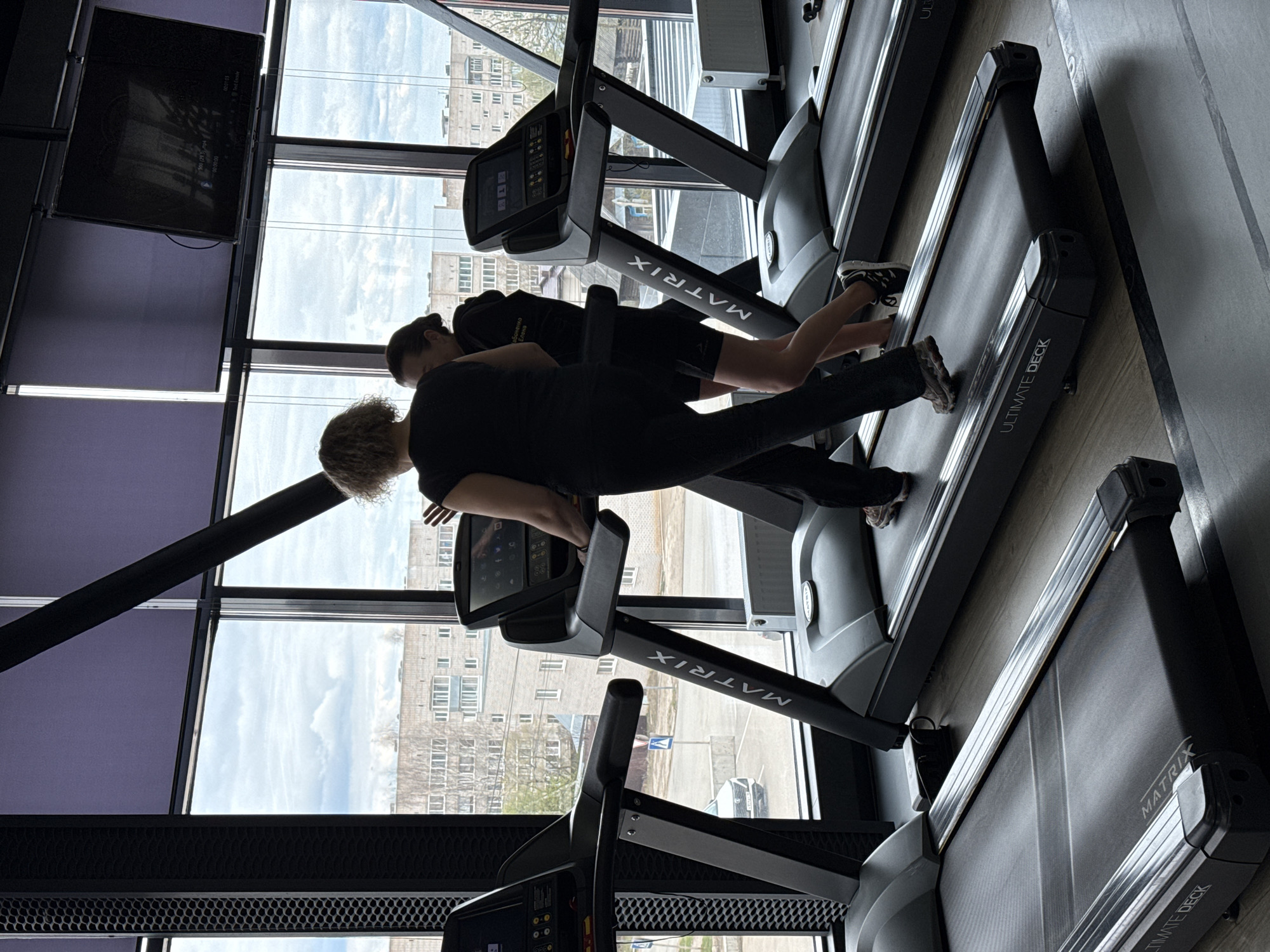 This image captures an indoor gym scene, specifically focusing on two individuals exercising on treadmills near large windows. The perspective is slightly elevated, looking down along the treadmills and out toward the urban environment beyond.
The primary subjects are two people, likely women, viewed from behind. In the foreground, a person with short, curly light-colored hair is seen walking or jogging on a treadmill, wearing dark attire. Behind them, another person with dark hair pulled back is also on a treadmill, wearing dark shorts and a dark hoodie.
Several treadmills are visible, including the two in use. The brand name "MATRIX" is clearly visible in white letters on the side supports of the treadmills, and "ULTIMATE DECK" is seen on the deck section of at least one machine. Each treadmill features a digital console screen. In the upper left of the frame, a flat-screen television is mounted, displaying what appears to be a video or media interface with a visible timestamp "00:21:03" and fragmented text like "Text_Movie".
The setting is a modern fitness center, characterized by dark equipment and structural elements. Large windows form a significant part of the wall, offering a view of buildings and a street outside, suggesting an urban or suburban location, consistent with Votkinsk, Russia. The sky outside is partly cloudy, indicating daytime. The individuals are actively engaged in cardio exercise.
On the back of the hoodie worn by the person in the background, some Cyrillic text is visible, although inverted relative to the wearer's position: "Зорошенко" and "ТВОЯ" (Tvoja/Your).