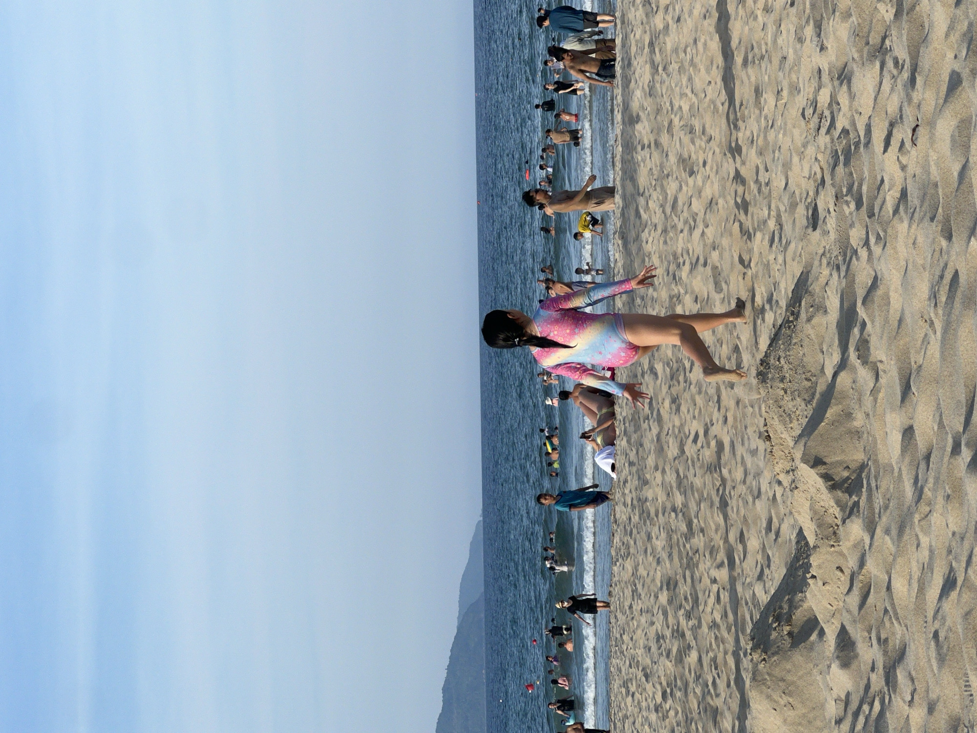 A group of people are enjoying a sunny day at the beach in Da Nang, Vietnam. The beach is sandy with some small sand dunes. The ocean is blue with gentle waves, and several people are swimming and wading in the water. In the foreground, a young girl in a colorful swimsuit walks on the sand, facing away from the camera. In the background, more people are visible in the water and on the beach. A distant coastline with mountains can be seen. The sky is clear and blue.