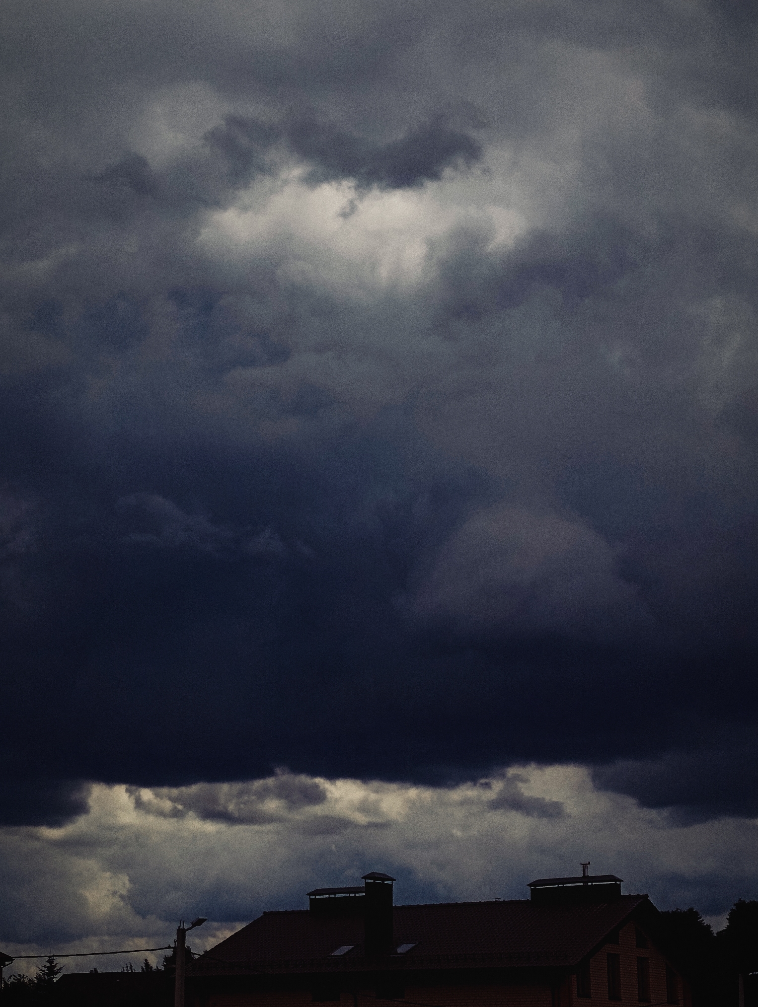 A low-angle, vertical outdoor photograph captures a dramatic sky filled with dark, heavy storm clouds. A bright, illuminated patch of sky is visible in the upper center, contrasting with the surrounding deep blues and grays. Below the turbulent sky, the rooftops of several buildings, likely houses, are silhouetted against the lighter clouds. The buildings appear to have red-tiled roofs and prominent chimneys. In the foreground, a lamppost stands to the left of center, and tree branches are visible on the far right. The overall atmosphere is moody and suggests an impending storm. The image is grainy, giving it a vintage or artistic quality. There are no people or animals visible. The location is specified as Minsk, Belarus.