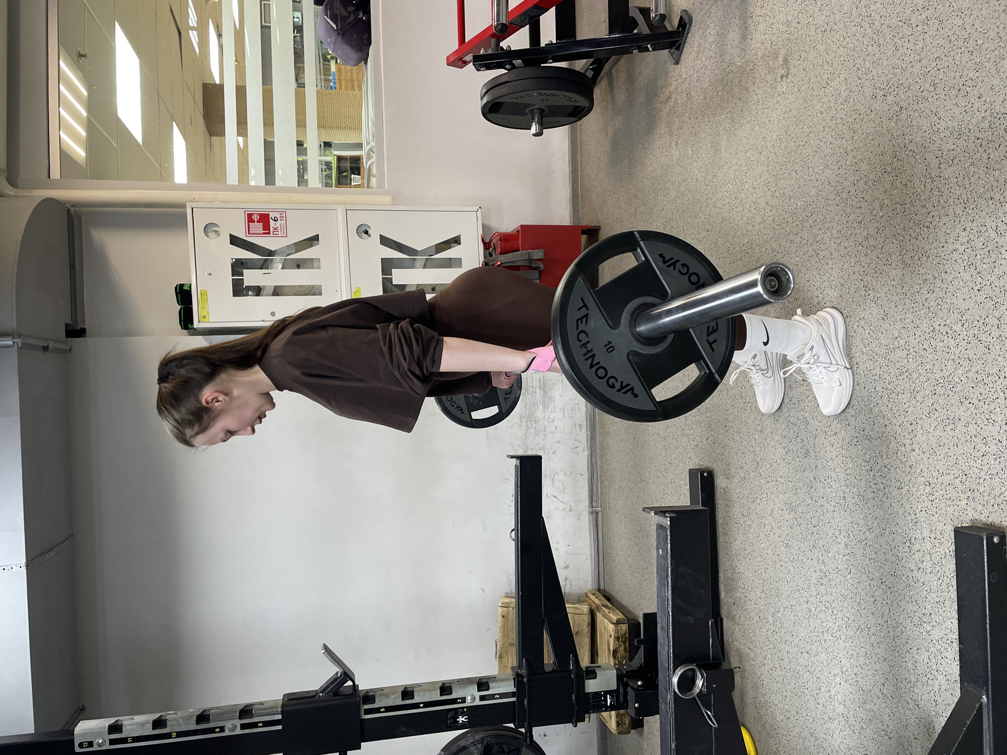 The image depicts a woman performing a bent-over row exercise in a gym setting. She is wearing a brown sweatshirt, brown leggings, white socks, and white sneakers. She is bent over, holding a barbell with weights on it.
The gym has light-colored walls and a speckled floor. In the background, there are fire safety equipment cabinets, a window, and other gym equipment. One of the weights on the barbell has "TECHNOGYM" and "10" printed on it.
The gym appears to be in Izhevsk, Russia, based on a sign on one of the fire safety cabinets.