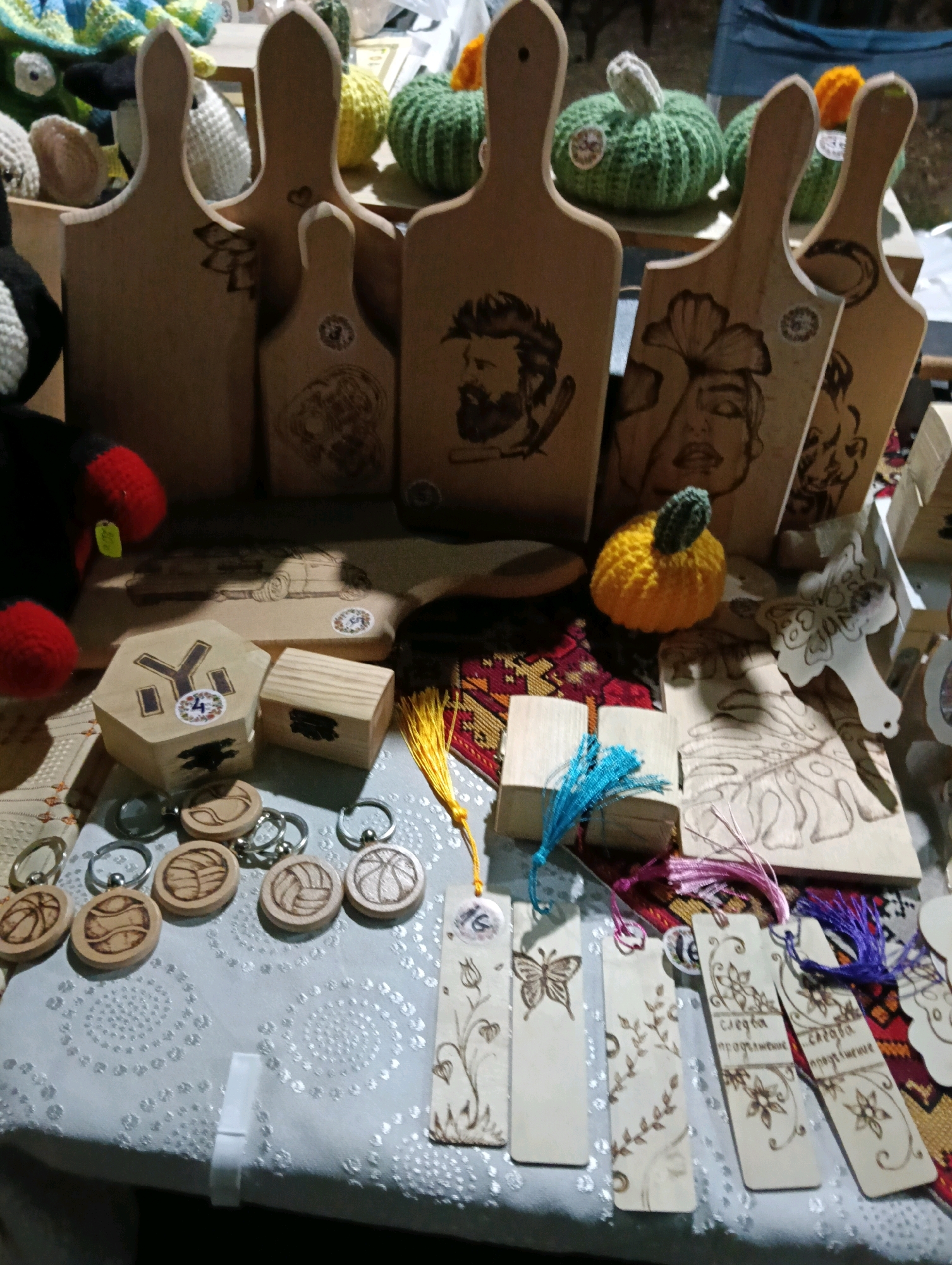 A display of handcrafted wooden items is presented, likely at an outdoor market or fair, given the natural lighting and the variety of handmade goods. The scene is shot from a slightly elevated angle, looking down at a table covered with a patterned white cloth.
Dominating the background are several large wooden cutting boards, each with a unique design wood-burned into its surface. These designs include portraits of men and women, as well as more abstract patterns. Behind the cutting boards, several crocheted pumpkins in green and orange, with green stems, add a festive touch. A dark stuffed animal with red accents is visible on the left.
In the foreground and midground, the table is laden with smaller wooden items. These include hexagonal and rectangular wooden boxes, some with decorative inlays or carvings. A collection of circular wooden keychains, each featuring a wood-burned sports-related design such as basketballs and volleyballs, are scattered across the cloth. Several bookmarks, also made of wood, are arranged in a row, each adorned with delicate wood-burned floral motifs and butterflies. Some of these bookmarks have colorful tassels attached. A small, yellow crocheted pumpkin with a green stem is placed among the wooden items. The overall impression is of a stall selling artisanal crafts. The location is identified as Unknown, Unknown.