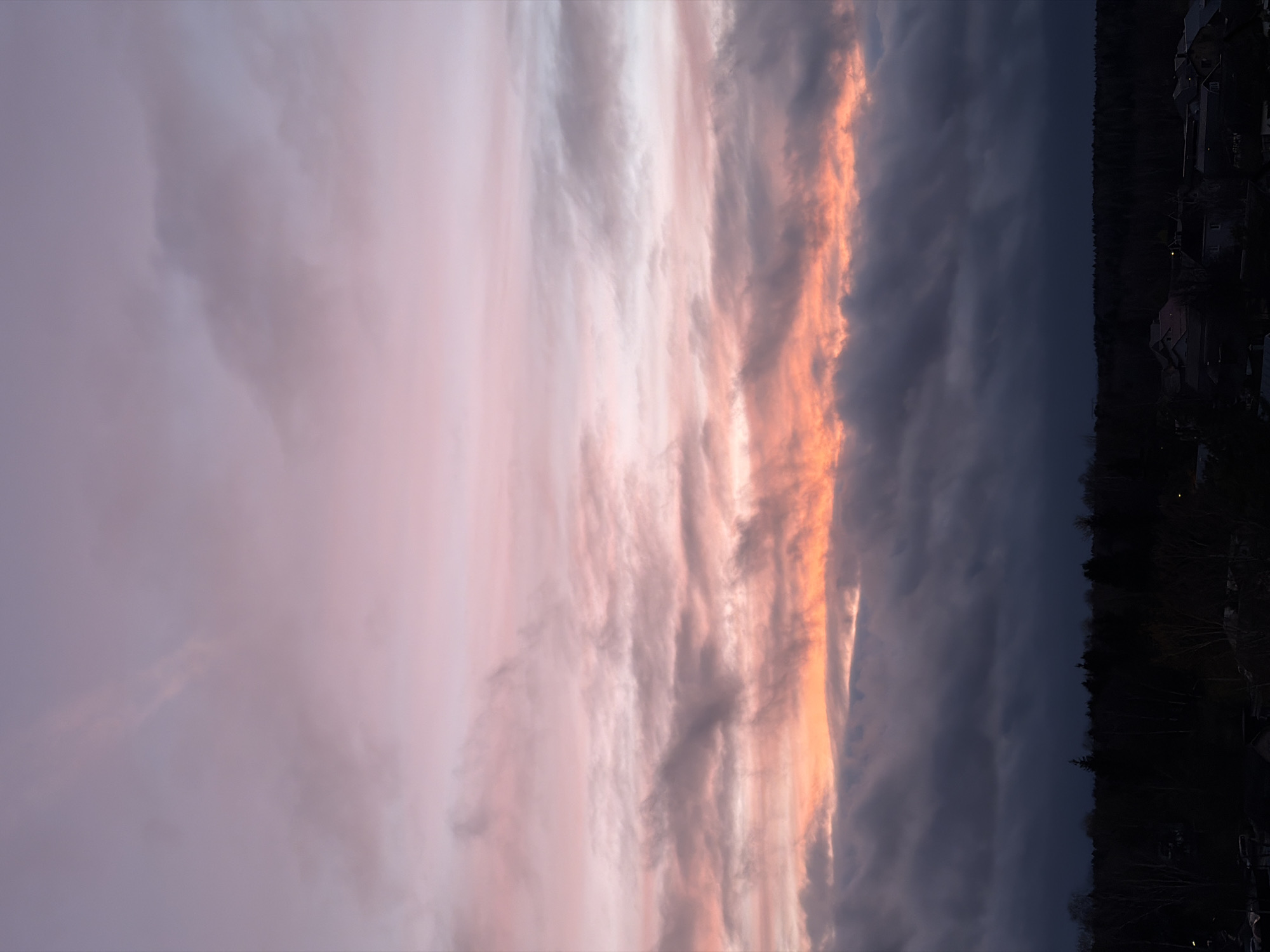 The image, presented vertically, captures a dramatic and colorful sky scene, likely at sunset or sunrise. The bottom portion of the image (when viewed horizontally) features a dark, silhouetted landscape. This landscape consists primarily of a dense treeline, suggesting a forest or wooded area, and scattered buildings or houses in the distance, some faintly showing small points of light.
The majority of the image is dominated by a vast and dynamic cloudscape. On the left side of the image (corresponding to the upper sky in a horizontal orientation), the clouds are softer and lighter, displaying gentle hues of pale pink, purple, and light grey, with diffuse and wispy formations. Towards the center, a prominent, elongated band of clouds stretches horizontally across the frame. These clouds are intensely illuminated with vibrant warm colors, including brilliant orange, golden yellow, and deeper pink, suggesting they are catching the direct light of the setting or rising sun. The textures within this bright band are varied, showing both smooth areas and more defined, streaky patterns.
Further to the right side of the image (corresponding to the lower sky), the clouds transition into darker, heavier masses of deep grey and blue, indicating areas that are already in shadow or moving further away from the sun's direct light. The overall scene emphasizes the natural beauty and striking contrast of the sky at dawn or dusk. There are no visible people, specific objects beyond clouds and distant structures, or discernible text in the image.