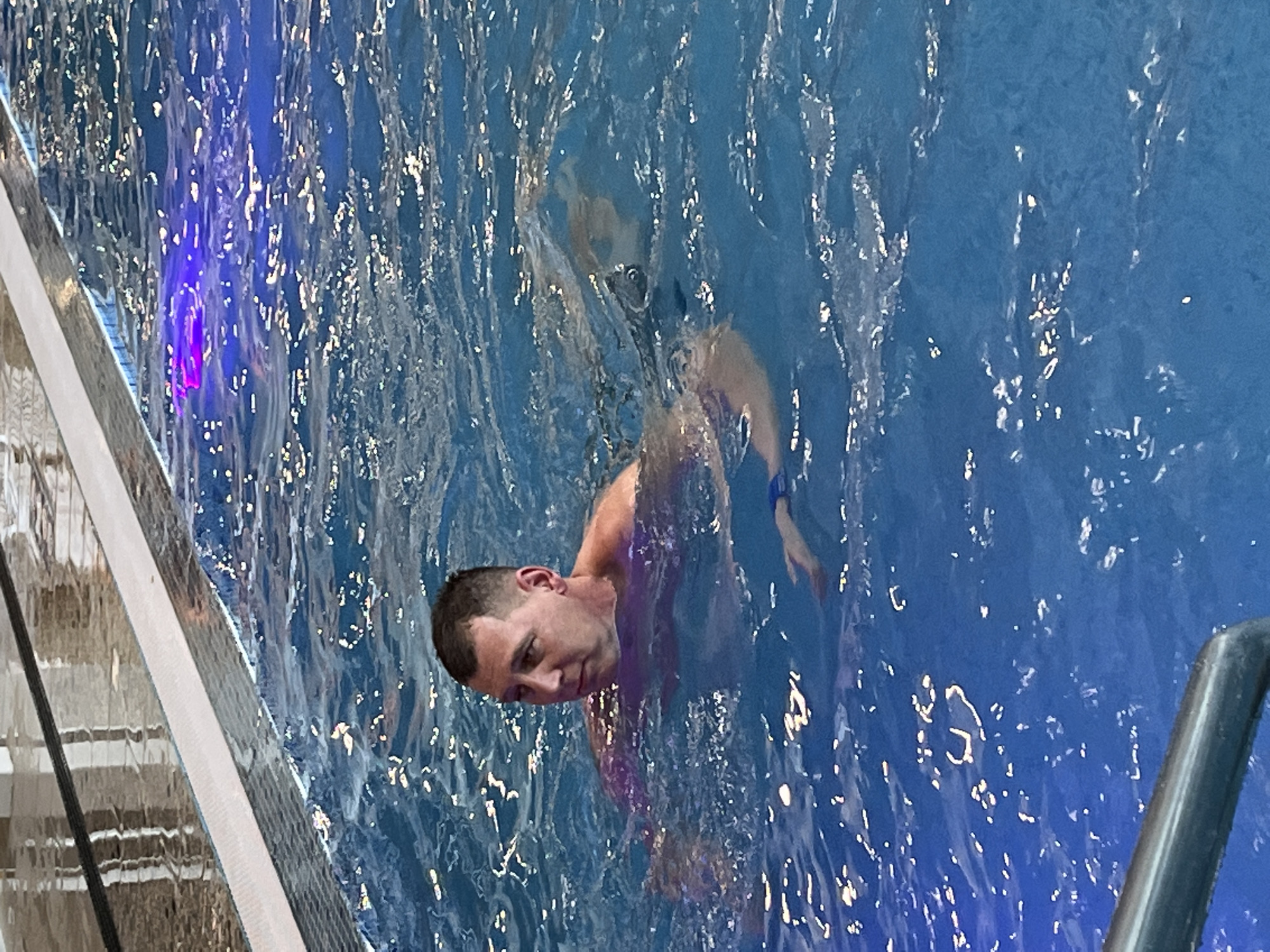 A man is swimming in a pool. The water is blue and reflective. There are ladders at the edge of the pool. There are no visible signs indicating the location is Votkinsk, Russia.
