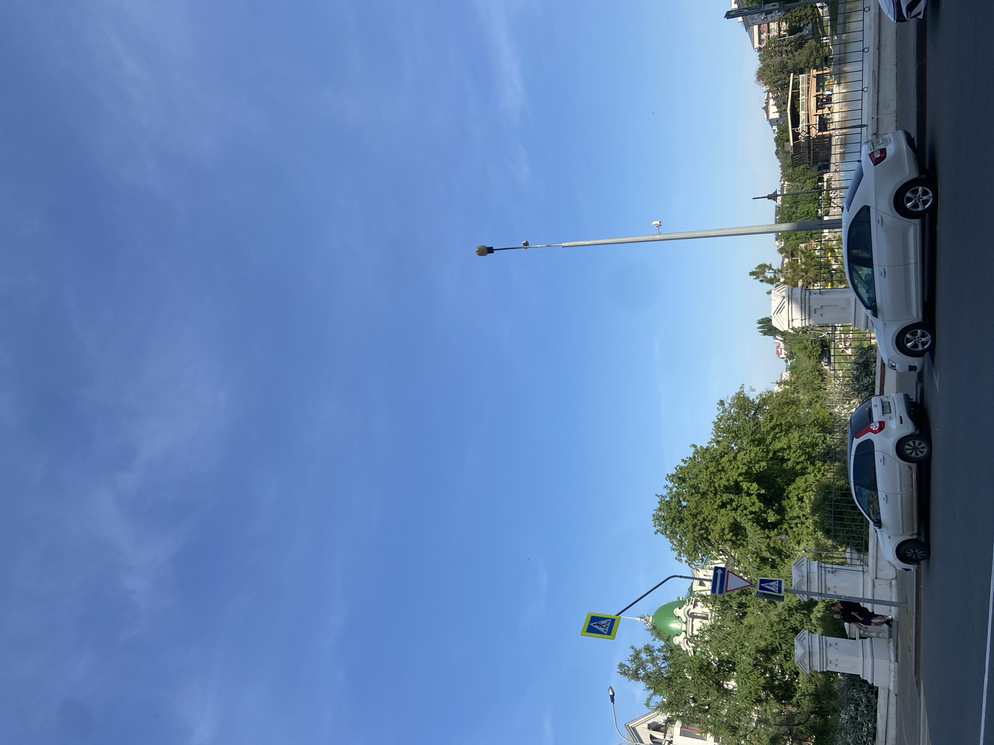 A daytime view of Kazan, Russia, shows a street scene alongside a waterway. Several white cars are parked along the roadside, near a low white stone barrier. Green trees line the road. Street signs are visible, including a yellow crosswalk sign and blue pedestrian signs. The background shows buildings with green domes. Overhead is a clear blue sky.